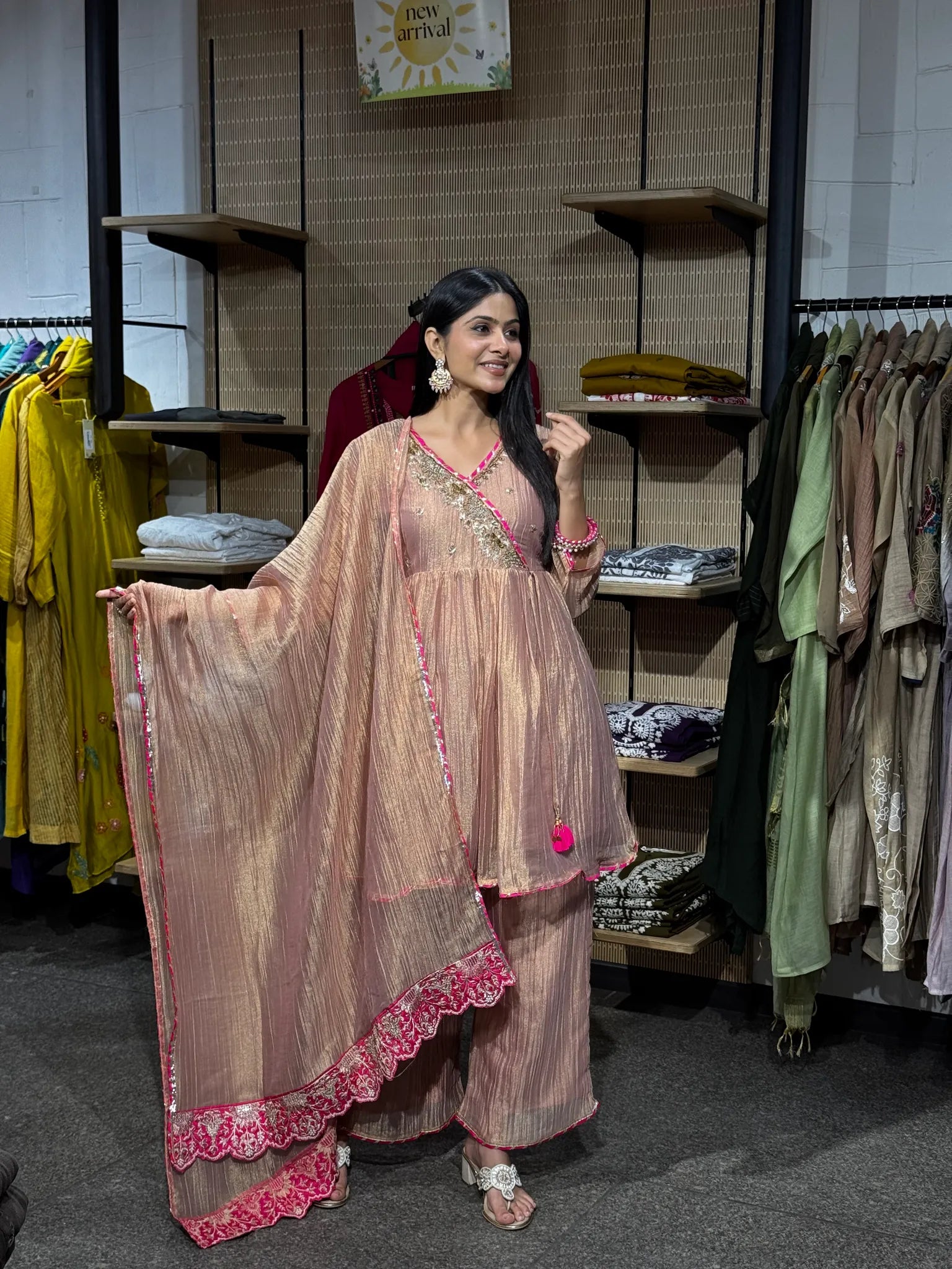 A woman modeling a baby pink tissue kediya sharara suit, holding the sheer embroidered dupatta open, inside a clothing boutique.