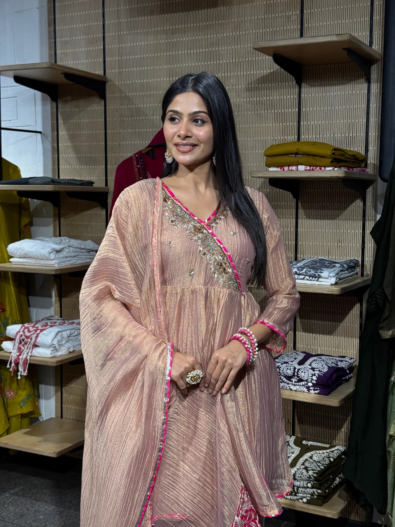 A woman wearing a baby pink tissue kediya sharara suit with a flared peplum top and wide sharara, inside a clothing store.