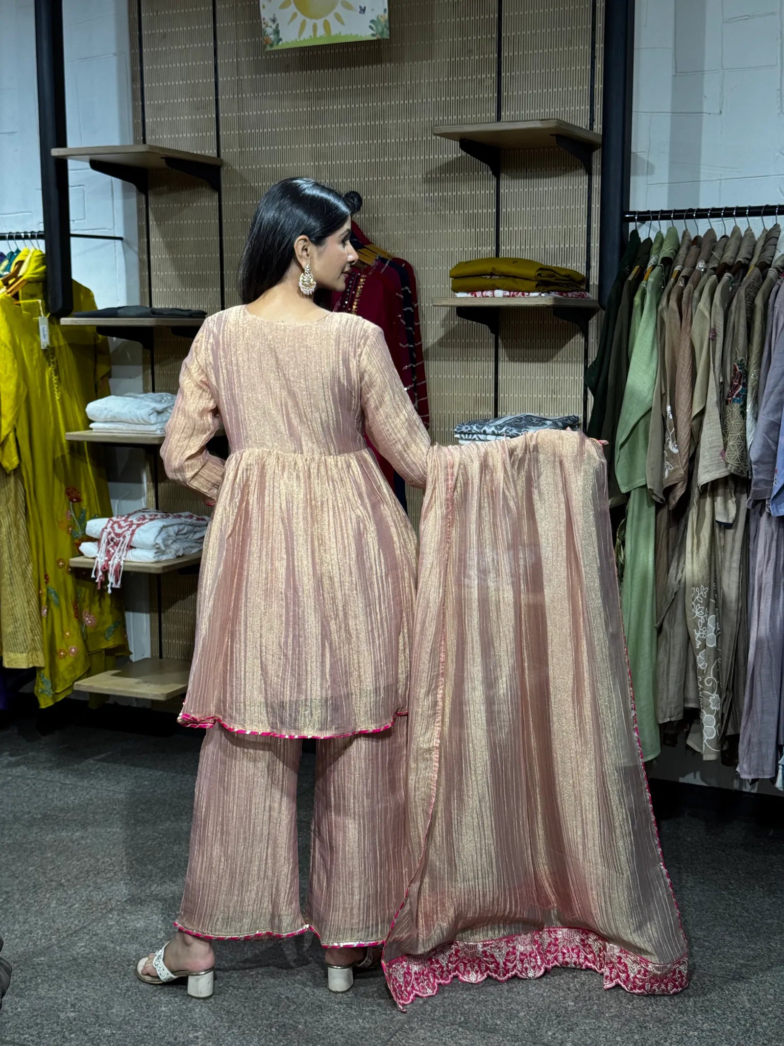Back view of a baby pink tissue kediya sharara suit, the model holding the dupatta open to show its embroidered scalloped hem.