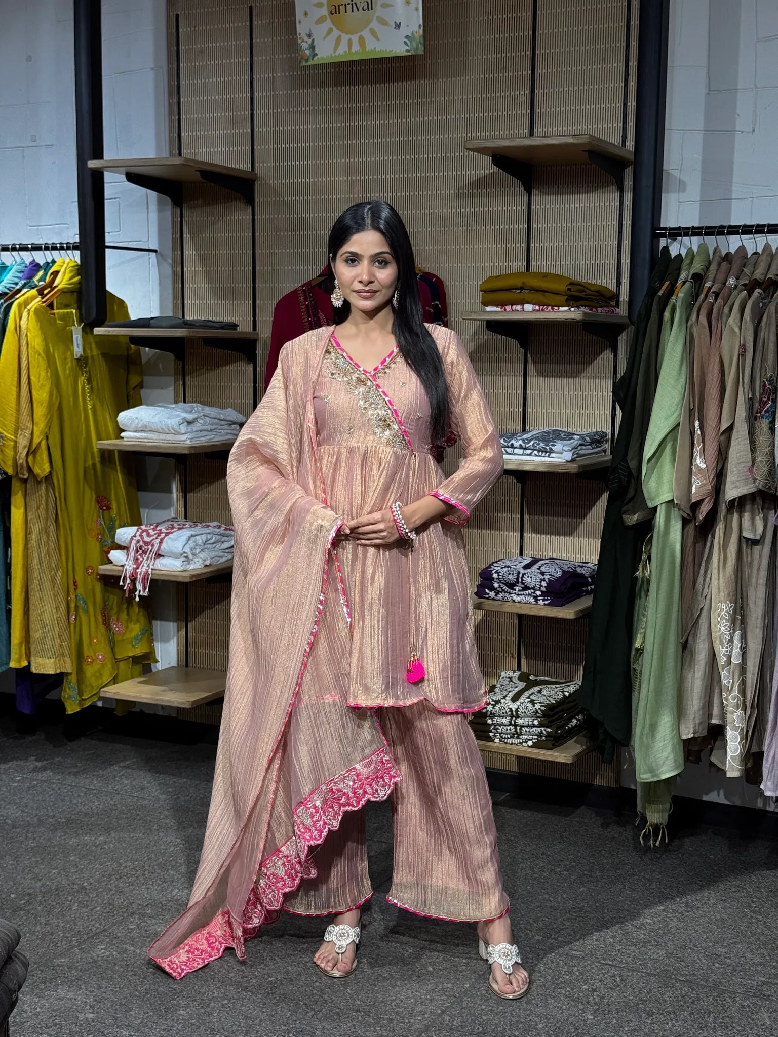 A woman modeling a baby pink tissue kediya sharara suit with the dupatta draped over one shoulder, standing in a boutique.