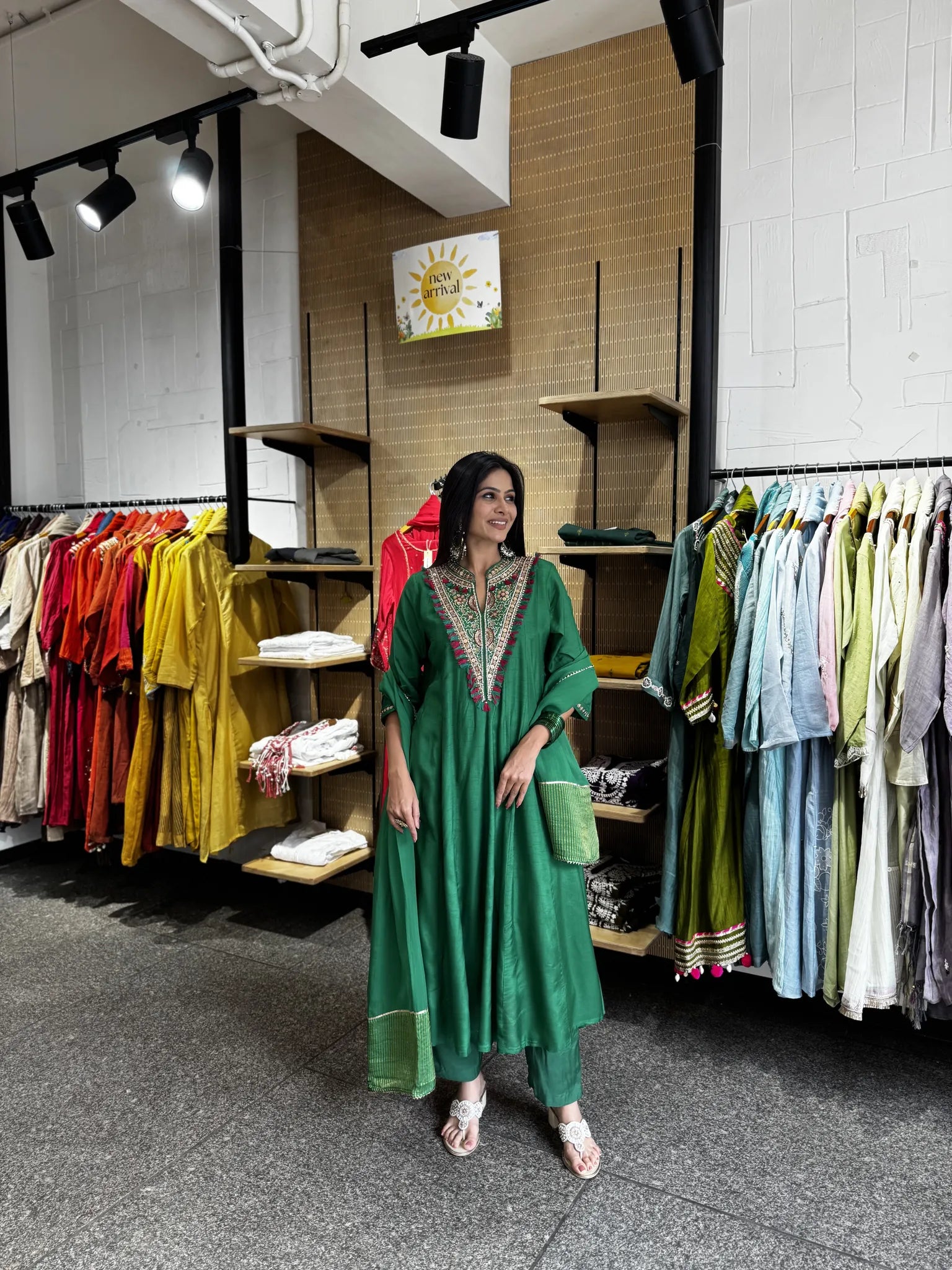 A woman wearing a green traditional outfit with embroidered neckline stands in a clothing store with racks of colorful garments in the background.