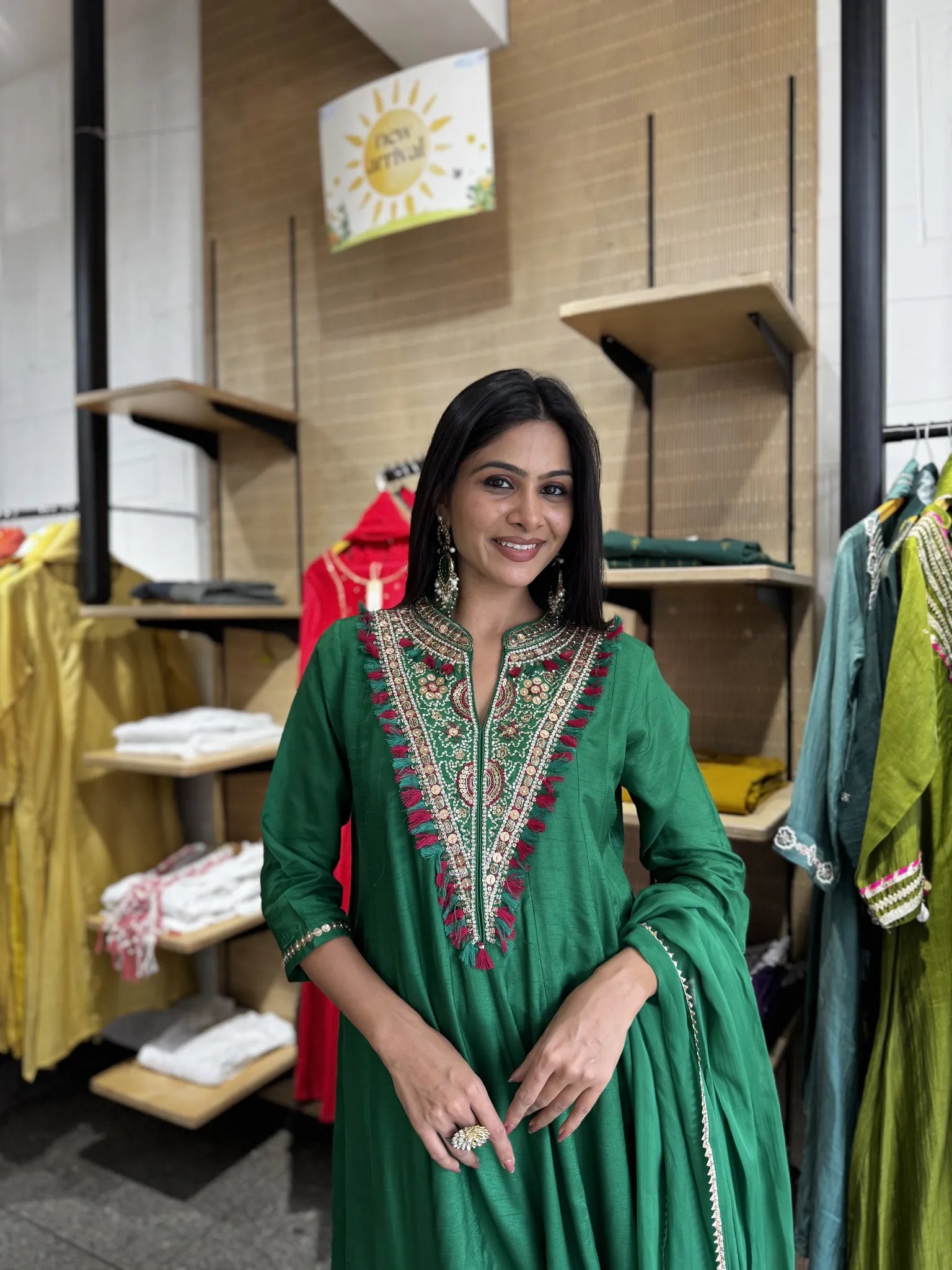 A woman wearing a green traditional kurta with intricate multicolor embroidery around the neckline, standing inside a clothing boutique.
