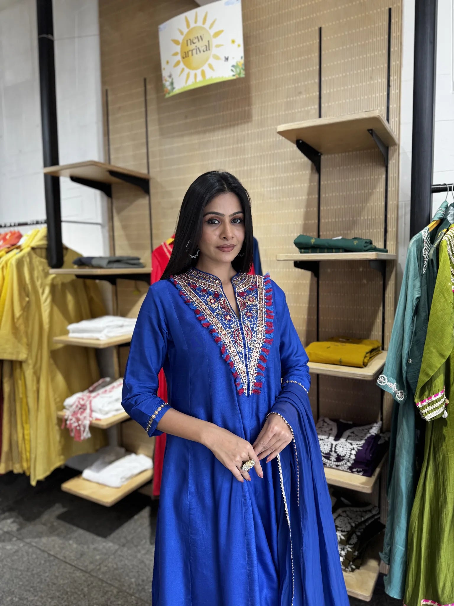 A woman modeling a royal blue Anarkali kurta with colorful embroidered neckline, standing inside a clothing store.