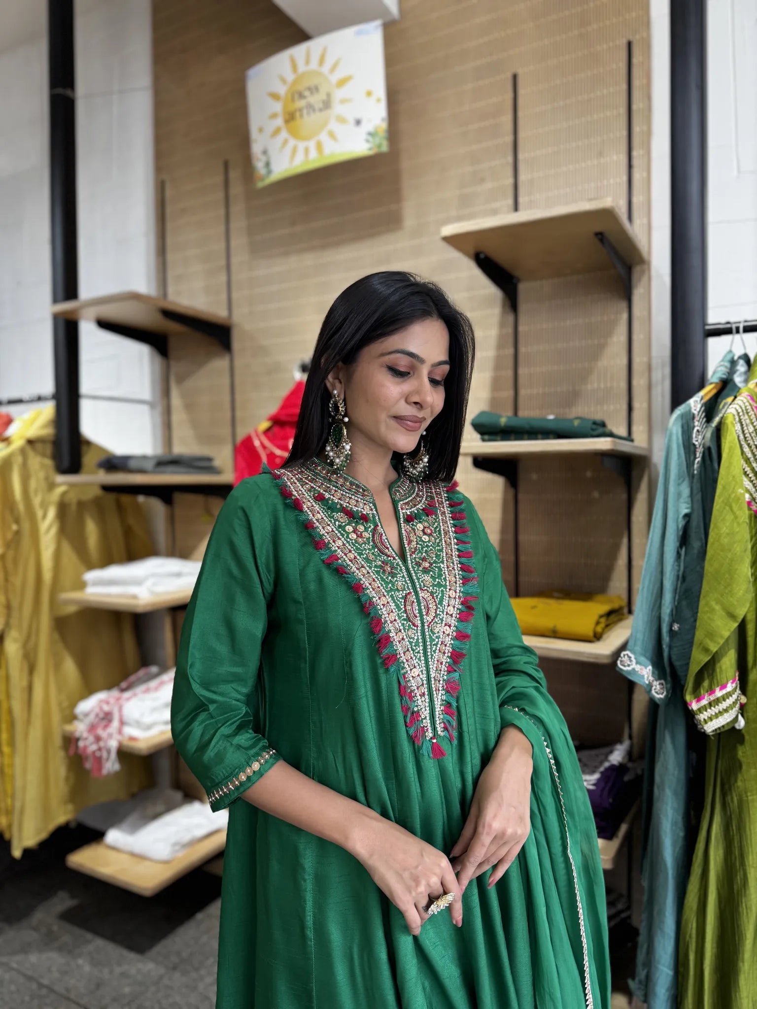 A woman wearing a green traditional embroidered kurta stands inside a clothing boutique. The kurta features a high neckline with intricate multicolor embroidery in red, gold, and white, and is paired with a matching green dupatta.
