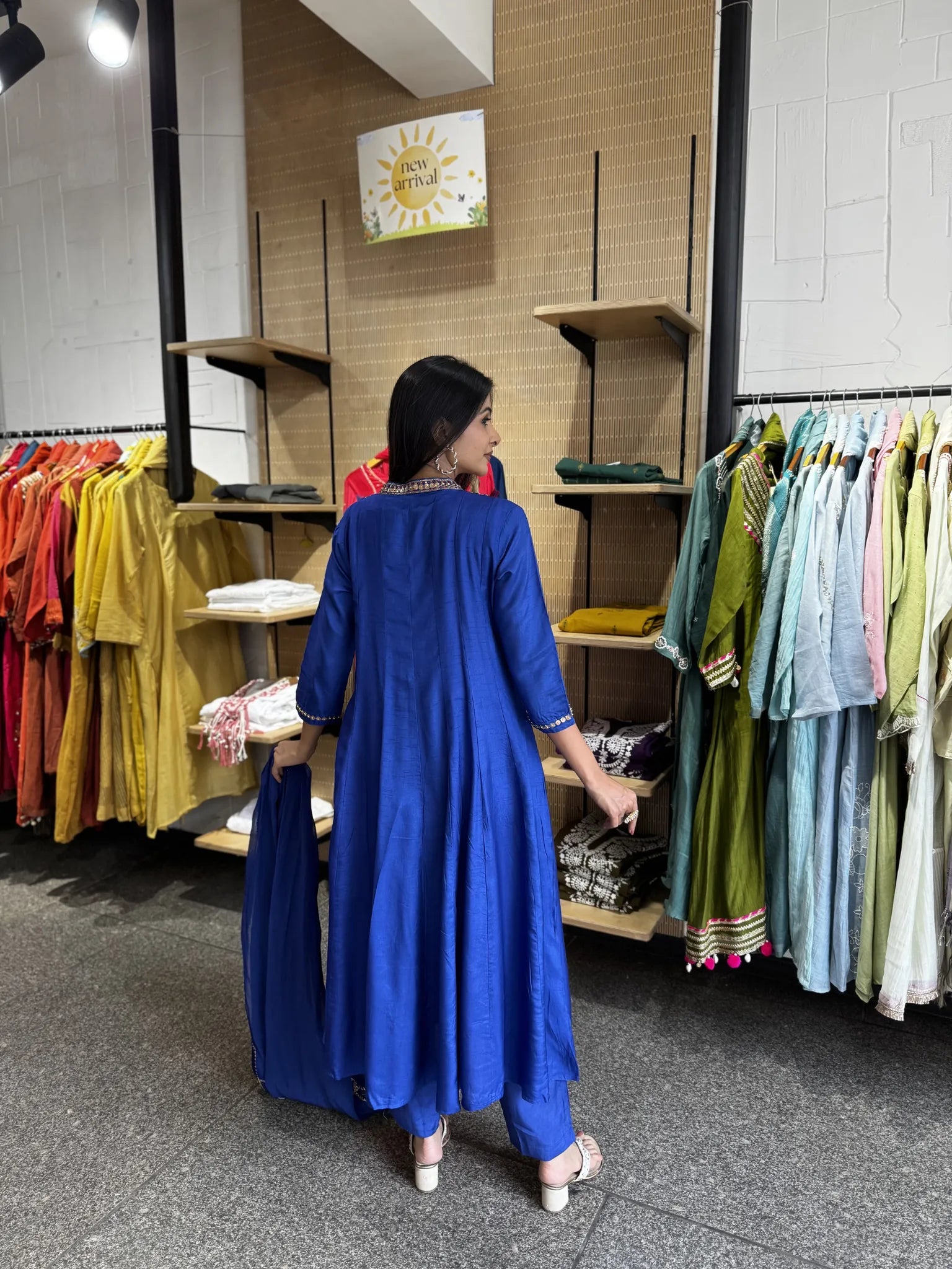 A woman wearing a bright royal blue kurta and matching trousers stands in a clothing store, holding a matching blue garment bag. The store has wooden shelves and racks filled with colorful clothing.