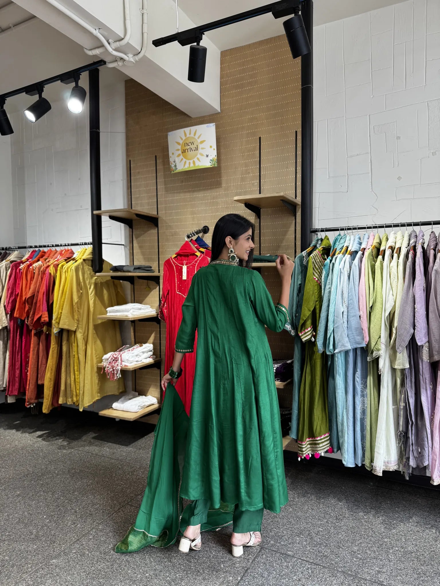 A woman wearing a green traditional outfit stands in a clothing store, looking at garments hanging on racks around her.