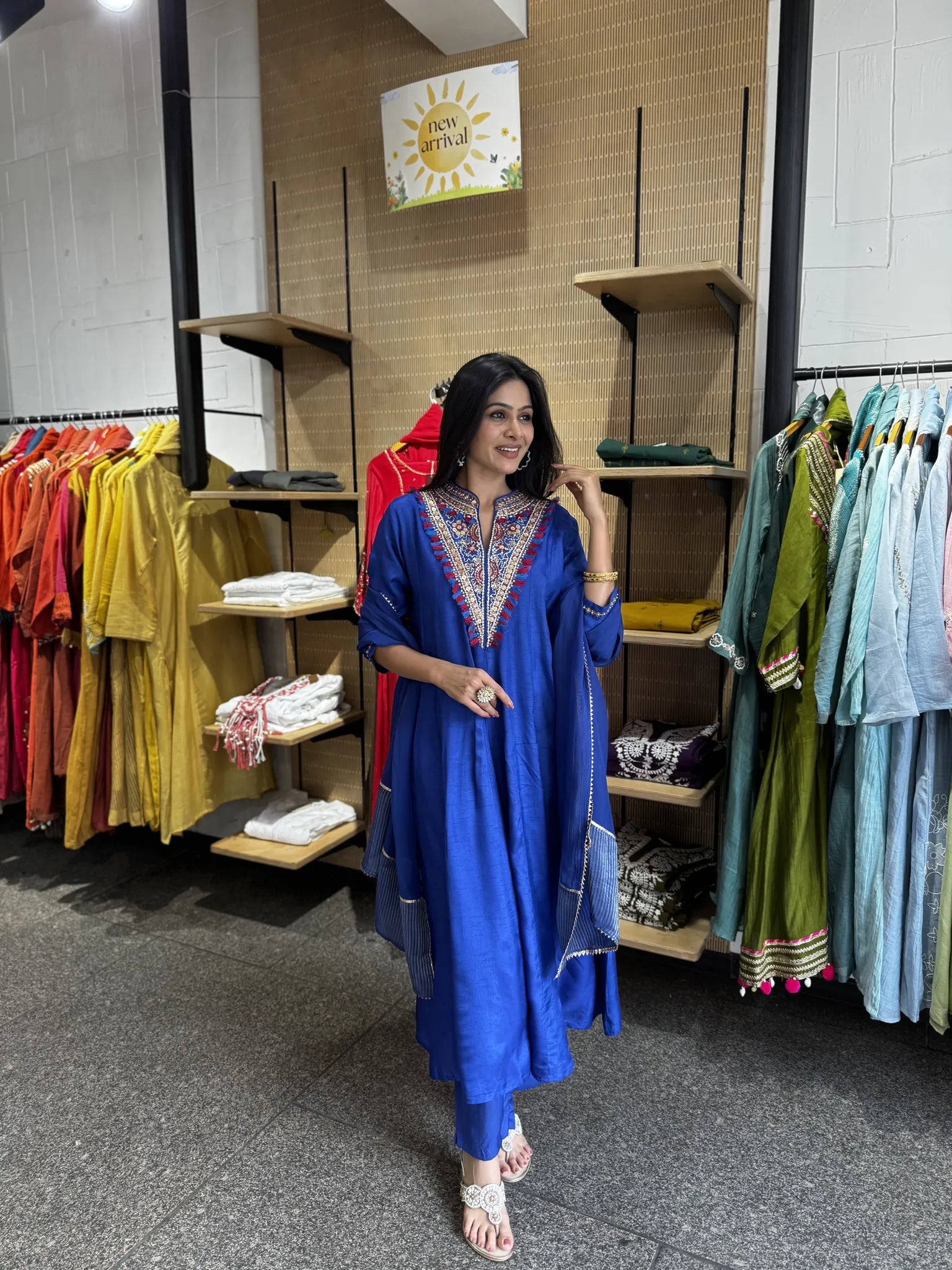 A woman modeling a royal blue kurta with colorful embroidered neckline and a matching sheer blue dupatta, standing inside a clothing boutique with racks of garments in the background.