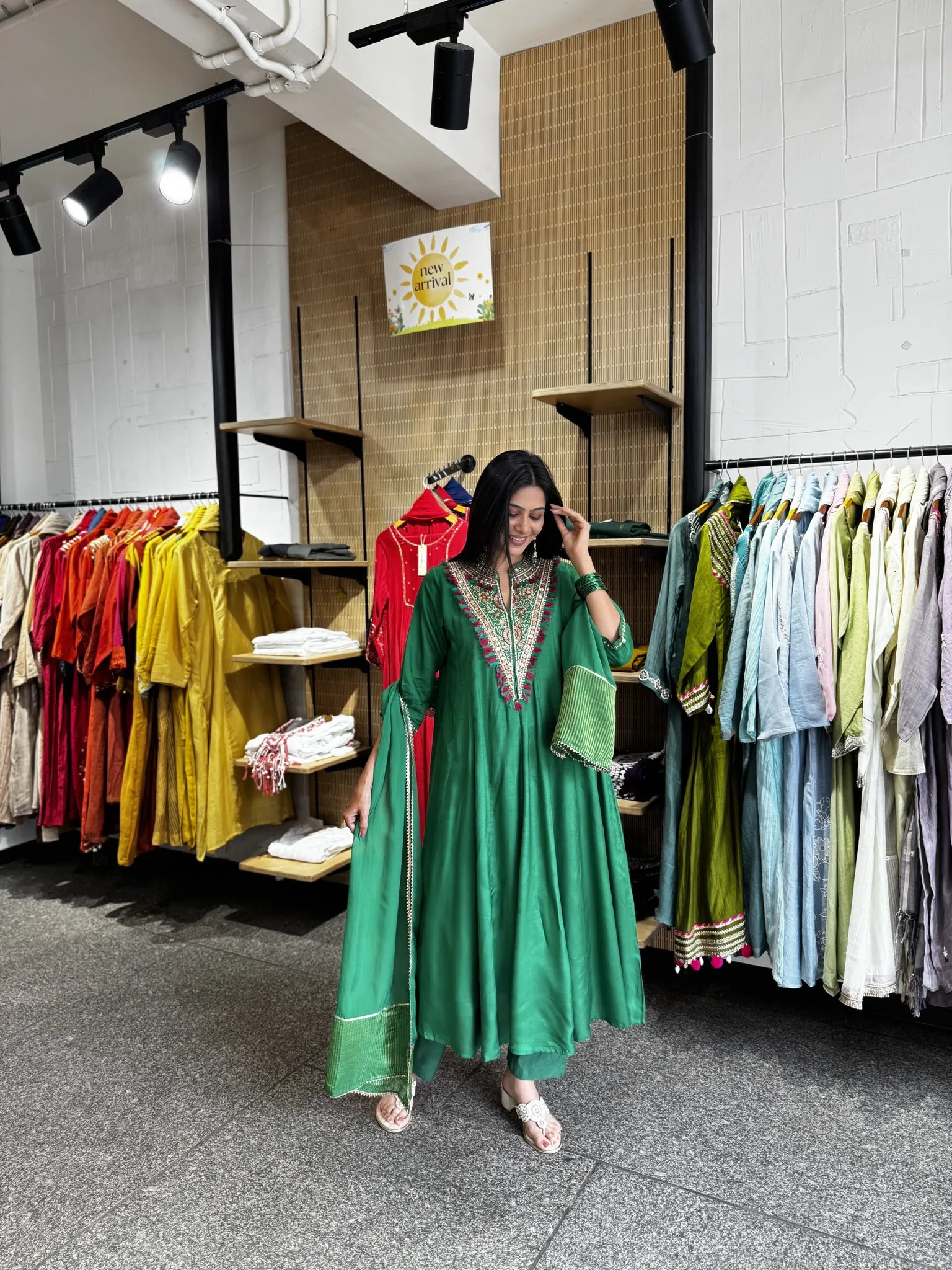 A woman wearing a green traditional kurta with embroidered neckline and a matching dupatta, standing inside a clothing boutique with racks of colorful garments in the background.