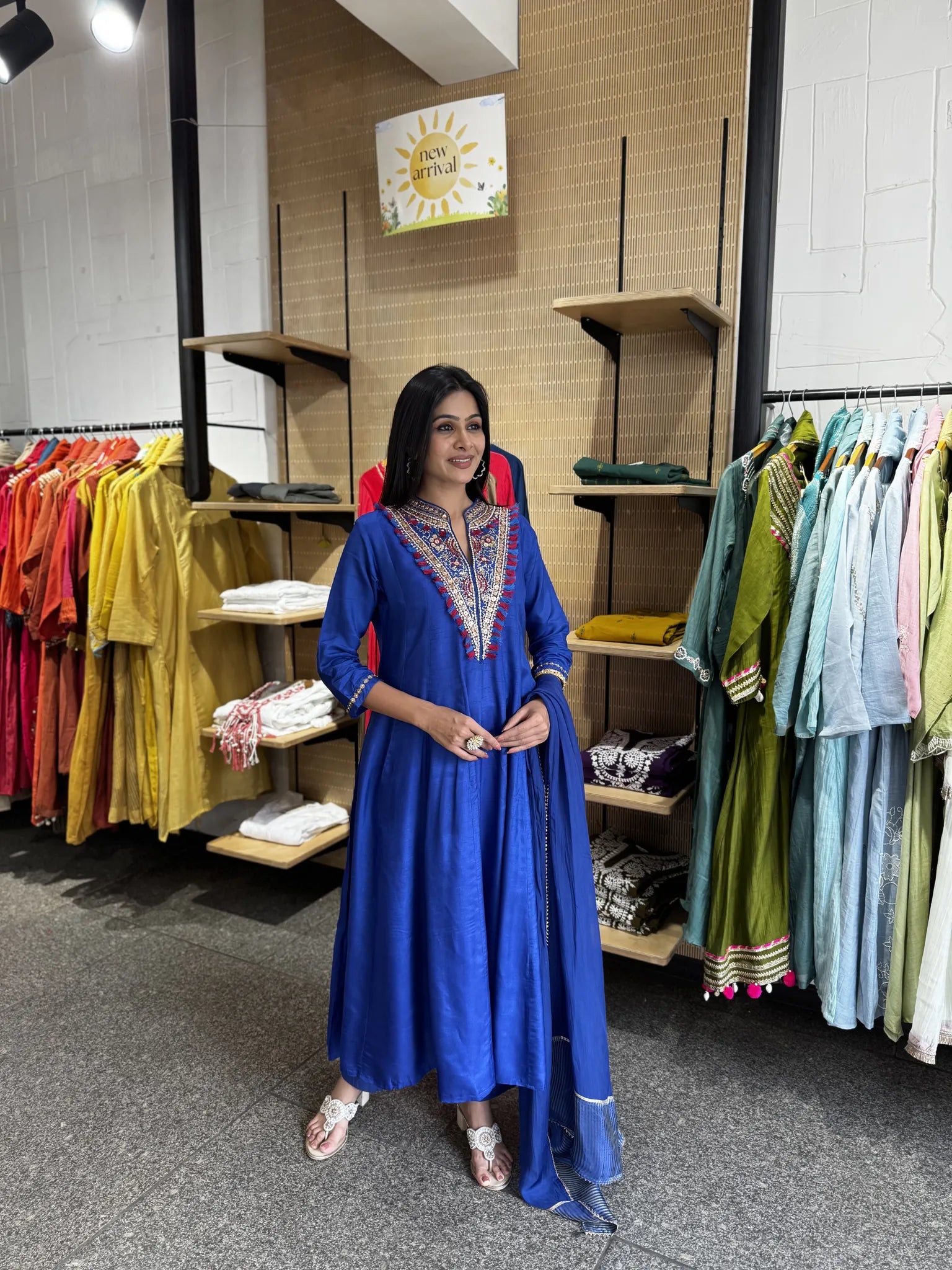 A woman modeling a royal blue traditional kurta set with embroidered neckline and dupatta inside a clothing boutique, surrounded by racks of colorful garments.