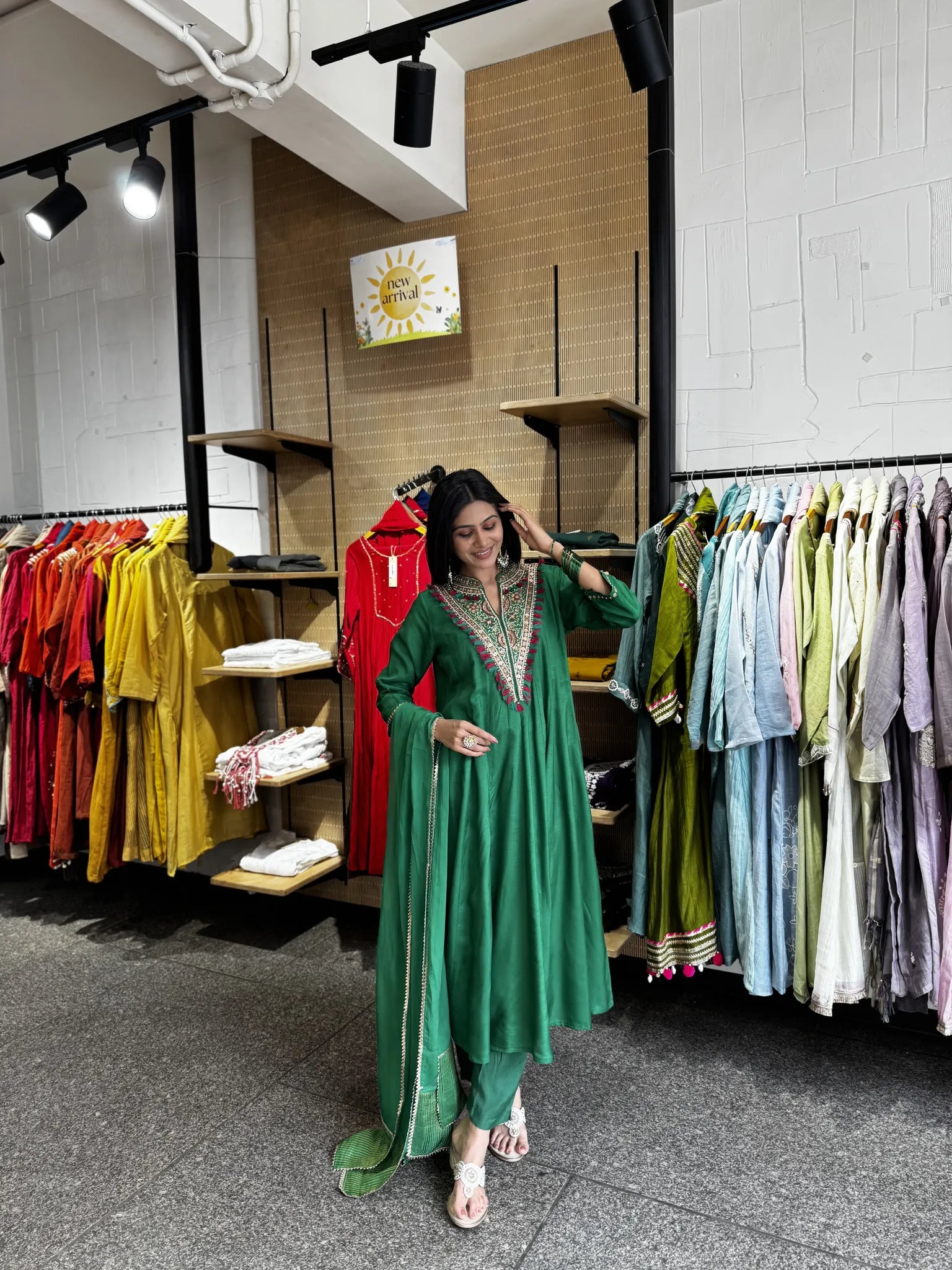 A woman modeling a green traditional outfit with silver embroidery in a clothing store, surrounded by racks of colorful garments.