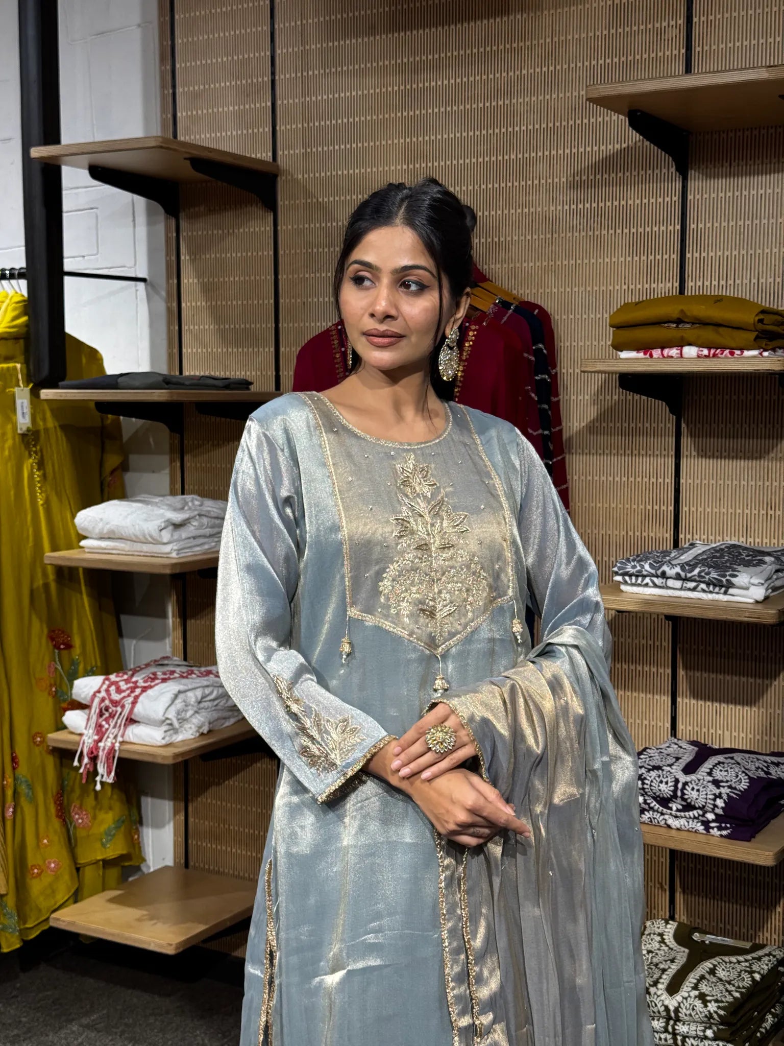 A woman wearing a pale blue tissue straight cut kurta with gold zari sleeve borders, hands clasped, inside a clothing store.