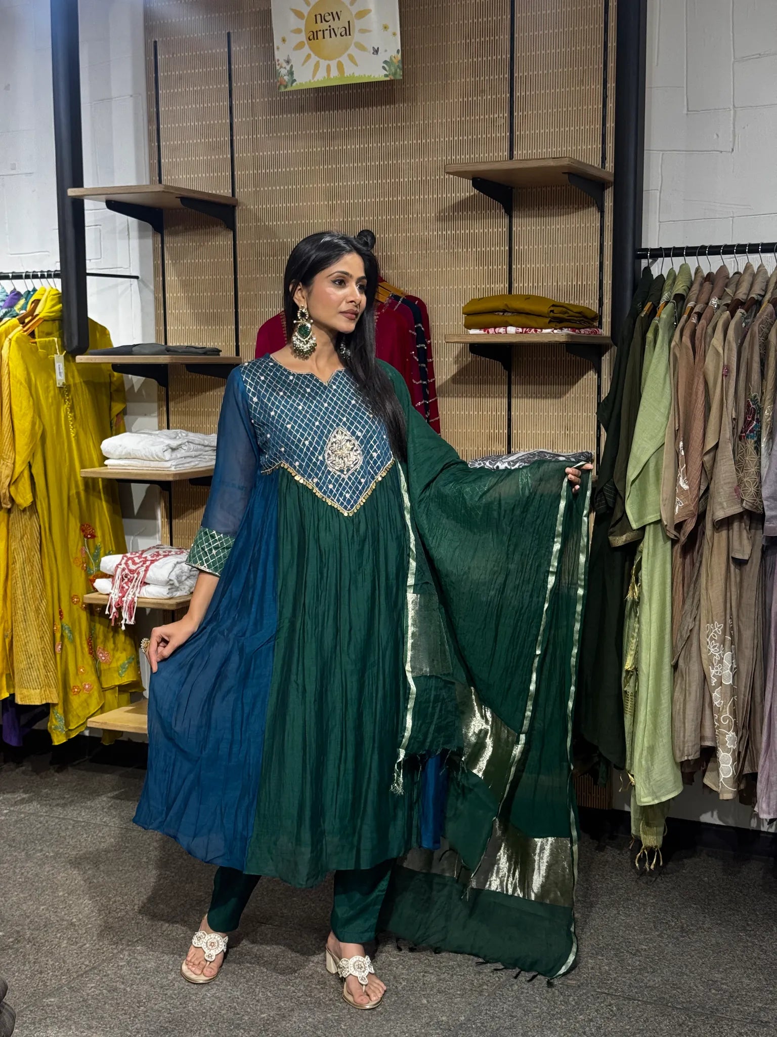 A woman modeling a teal and green mul chanderi A-line kurta pant set, holding the deep green dupatta open, inside a clothing boutique.
