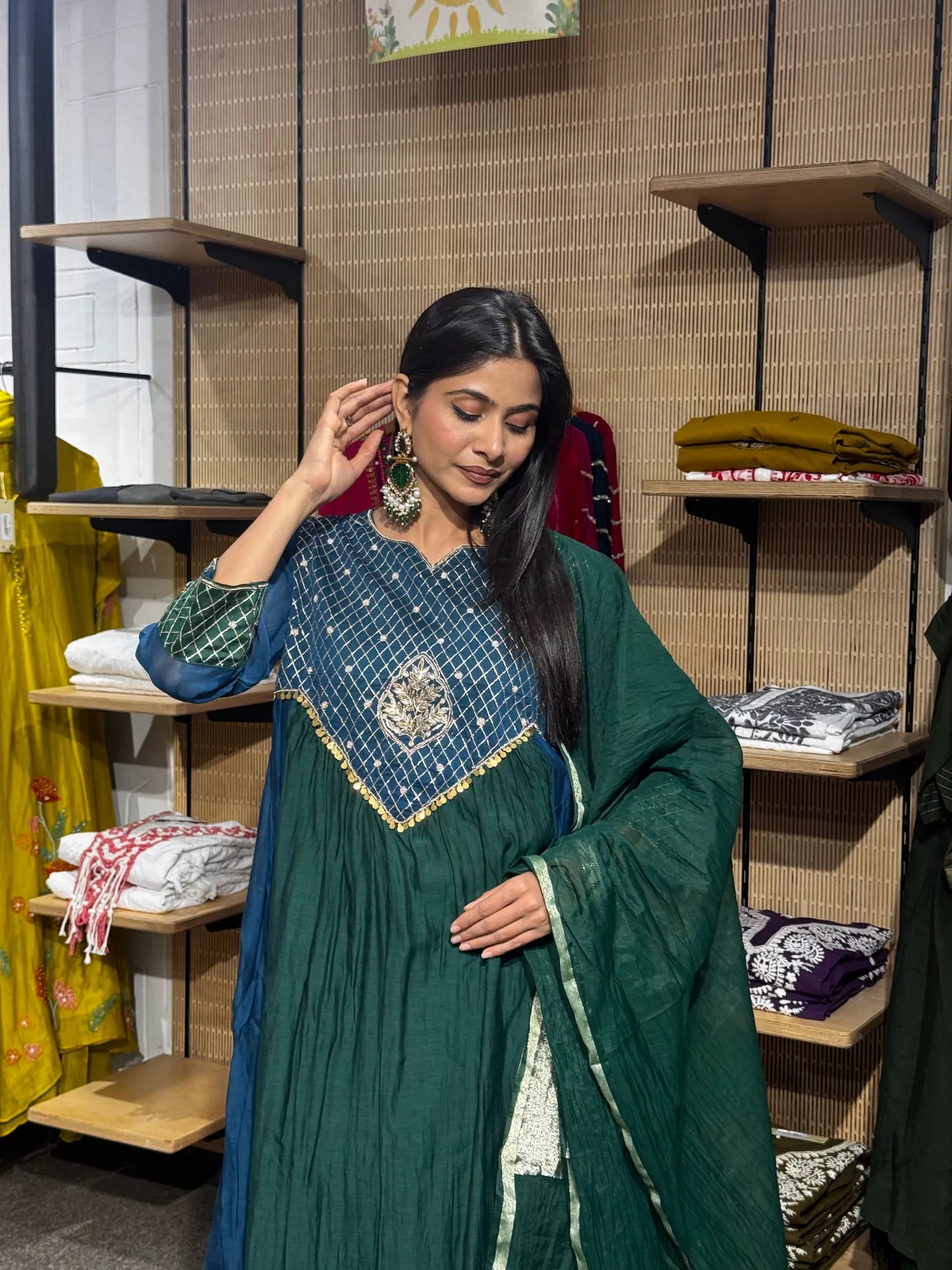 A woman wearing a teal green mul chanderi A-line kurta with a gold embroidered yoke, one hand raised, inside a clothing store.