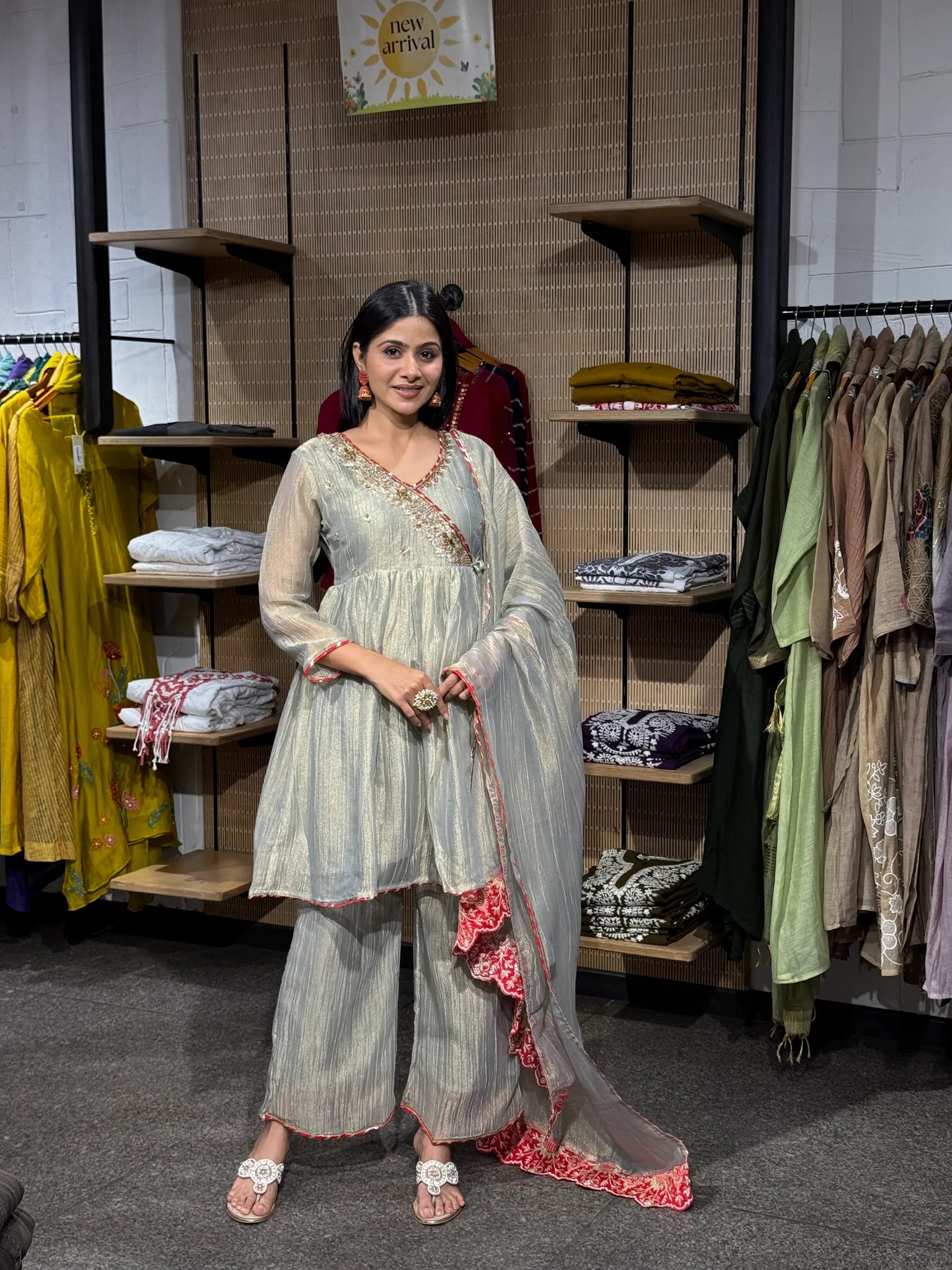 A woman modeling a cream tissue kediya sharara suit with red embroidered borders and a sheer dupatta, inside a clothing boutique.