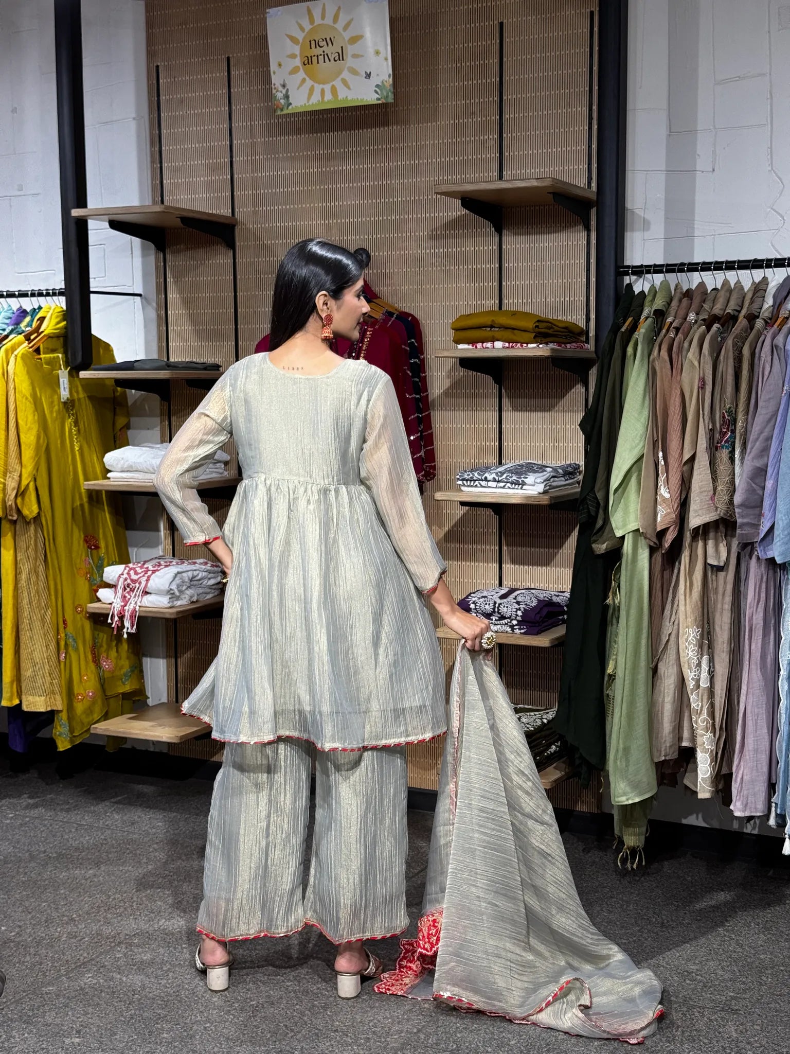 Back view of a cream tissue kediya sharara suit, the model holding the sheer dupatta open to show its red scalloped edge.