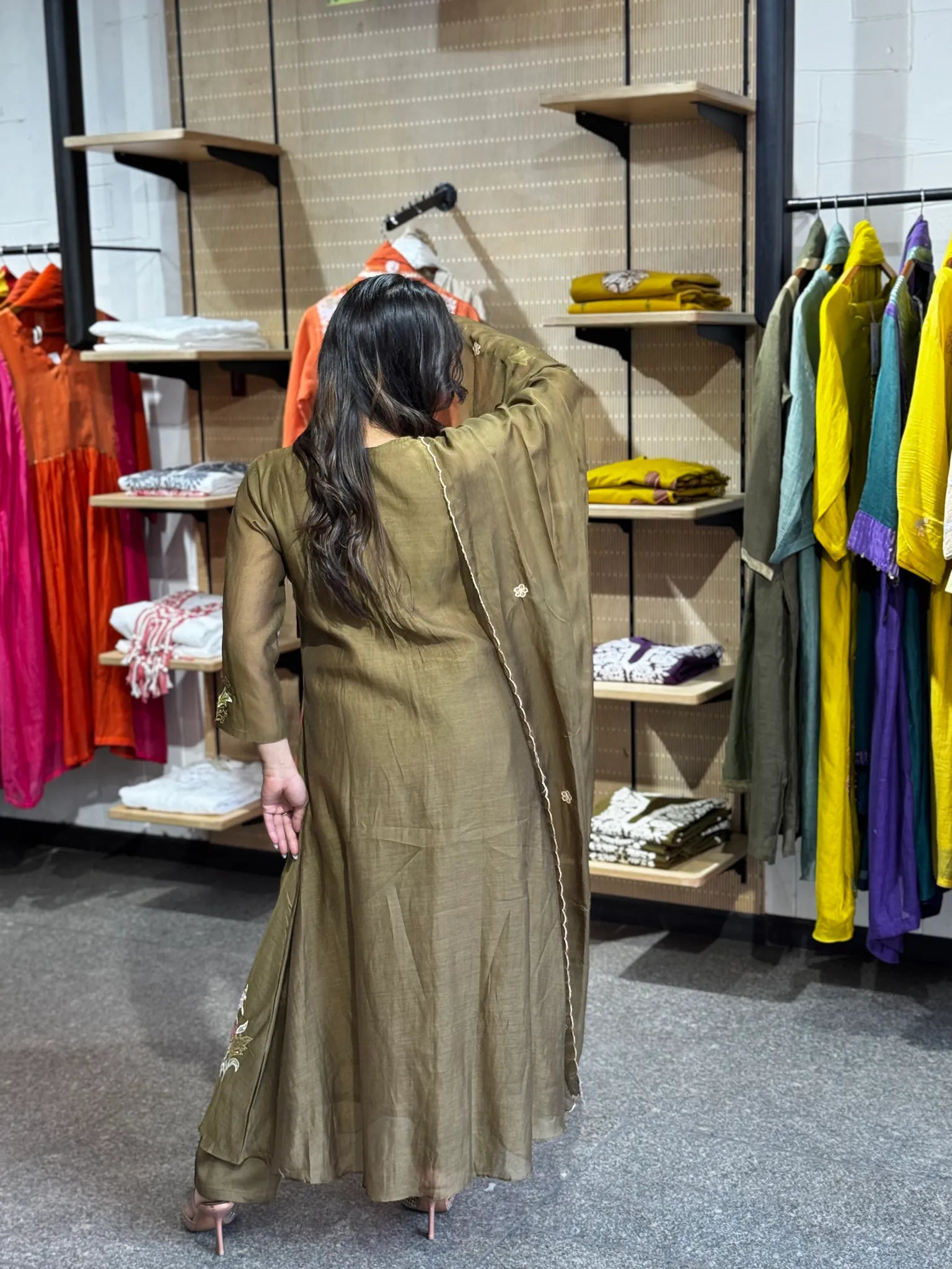Back view of the olive kurta set, the dupatta falling straight down the spine against rails of bright boutique clothing.