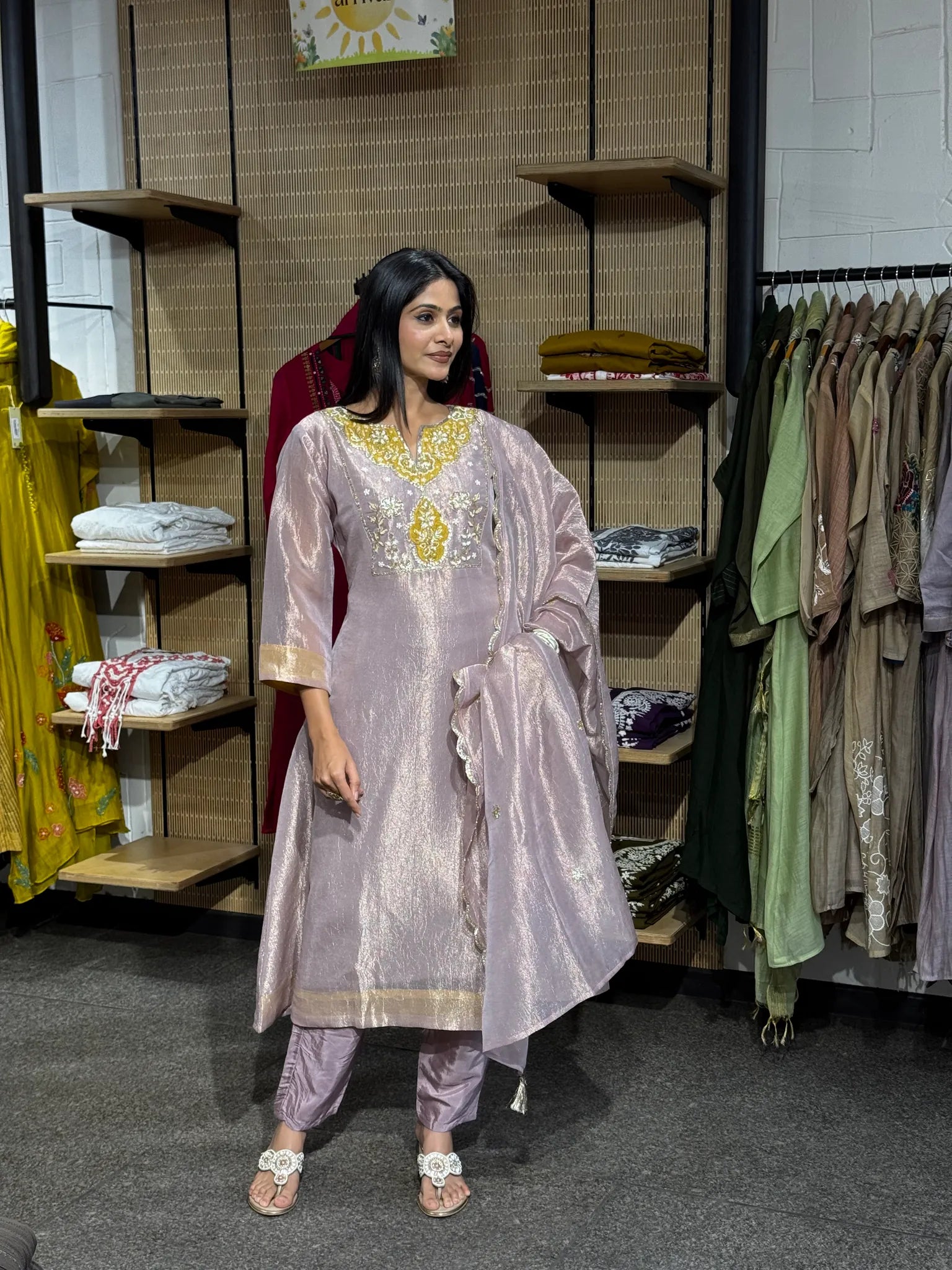 A woman modeling a lilac tissue A-line kurta pant set with a matching dupatta, standing inside a clothing boutique with racks of colourful garments.