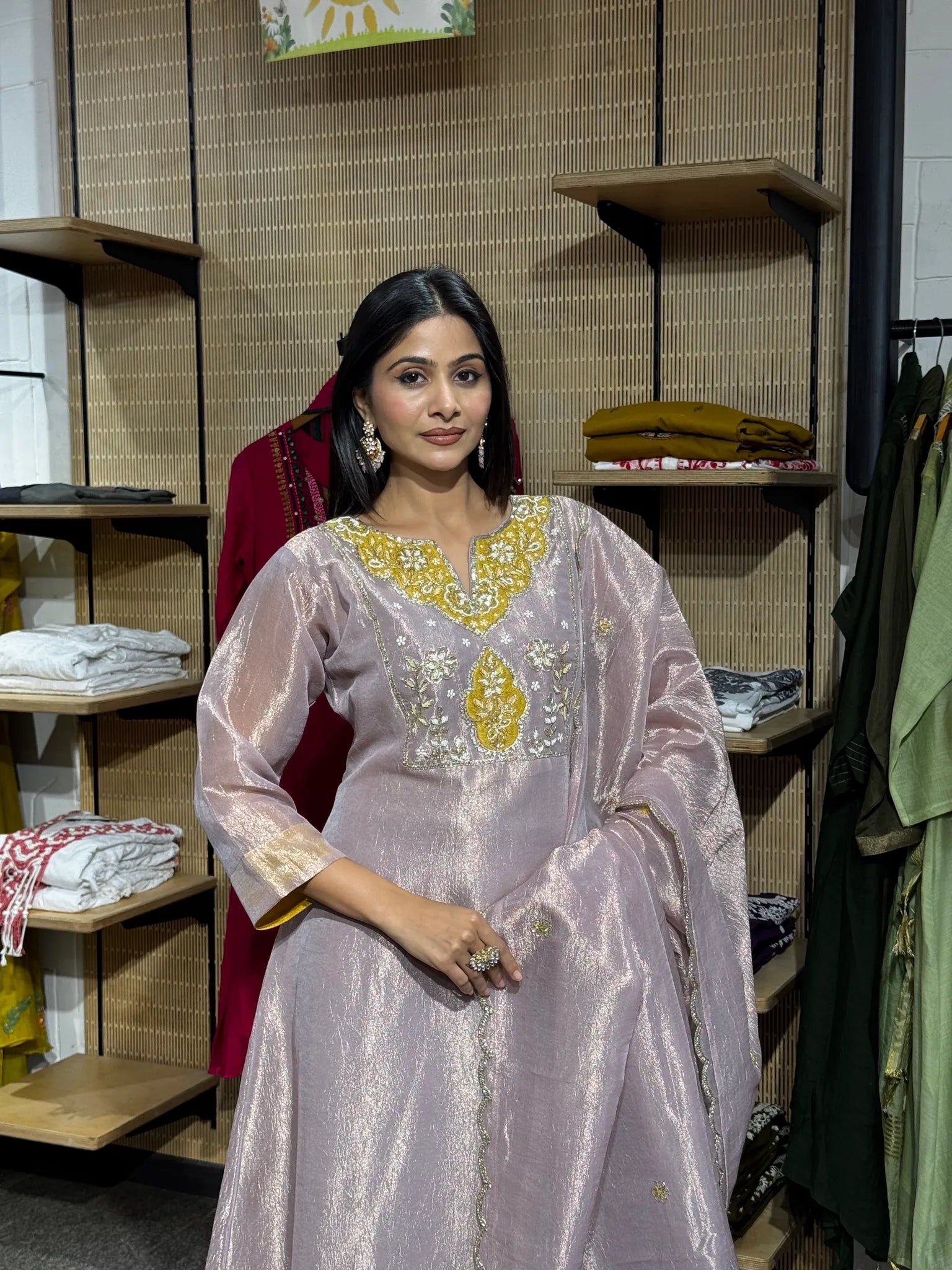 A woman wearing a lilac tissue A-line kurta with gold zari sleeve borders and an embroidered yoke, standing in a clothing store.