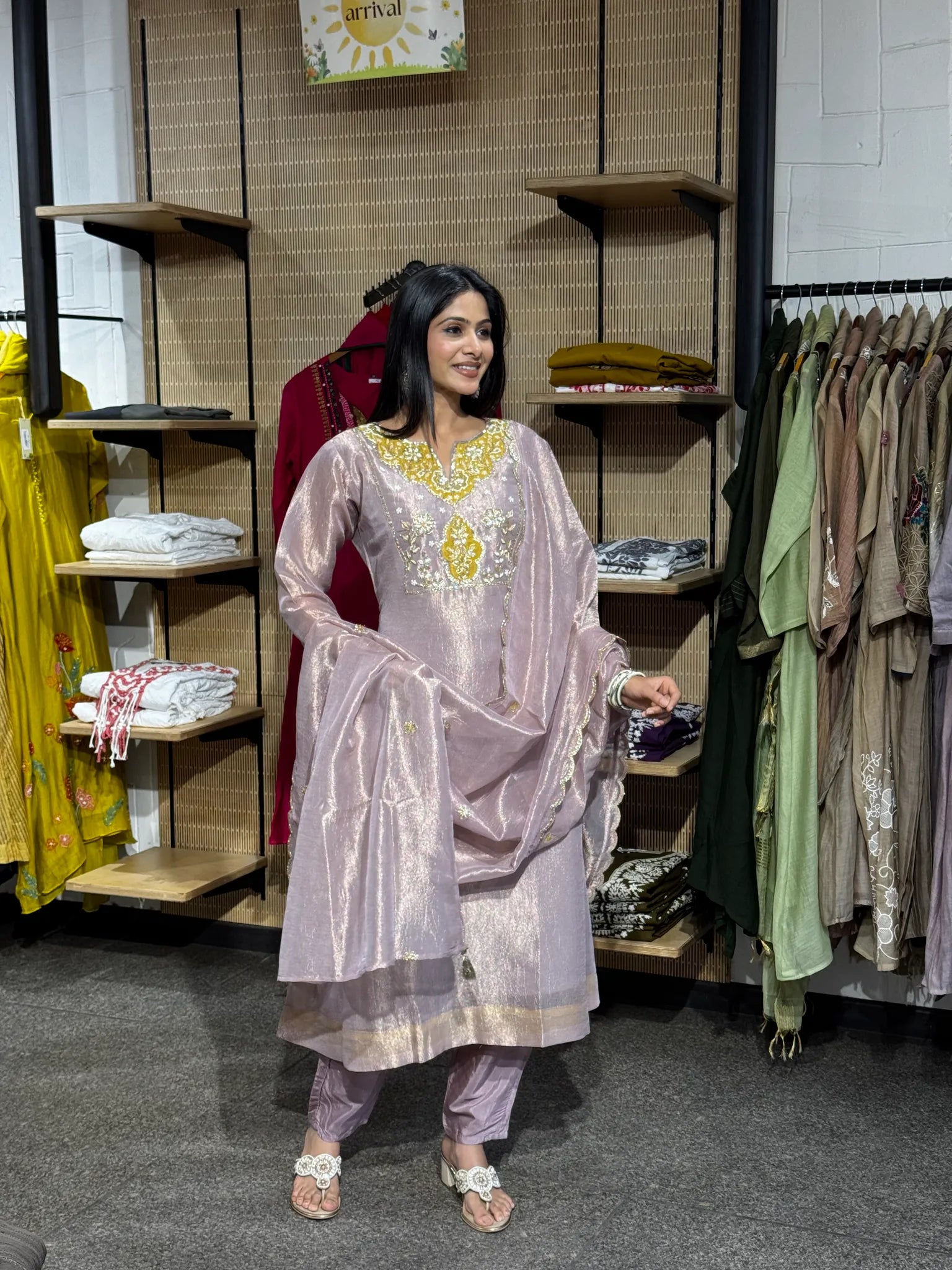 A woman modeling a lilac tissue A-line kurta pant set with the dupatta draped over one shoulder, inside a boutique.
