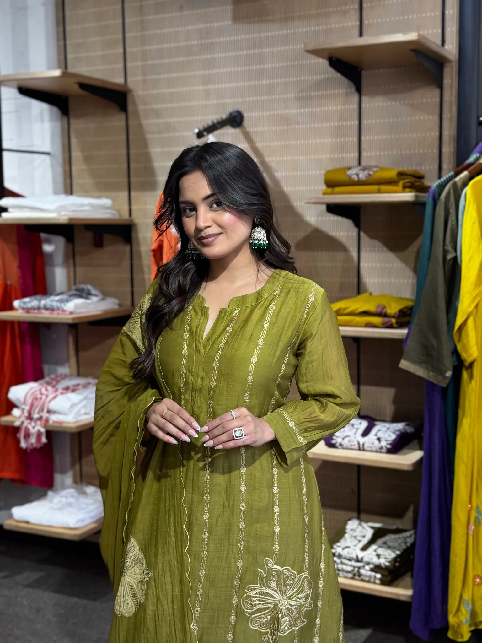 Waist-up in mehendi green, hands clasped, with a large cream flower embroidered low on the front of the kurta.