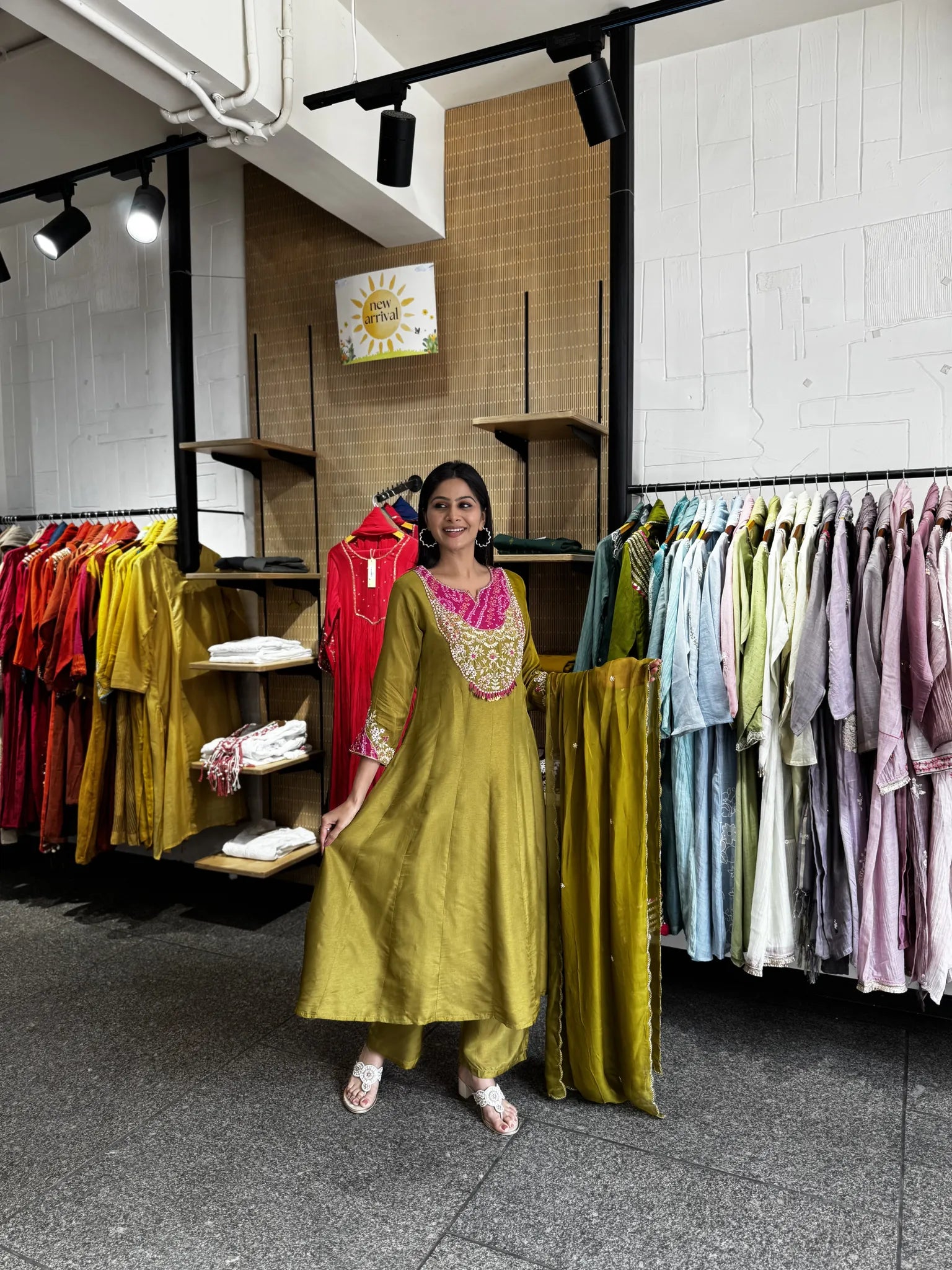 A woman modeling a mustard yellow-green kurta with pink embroidery and gold zari borders, standing inside a clothing boutique surrounded by racks of colorful garments.
