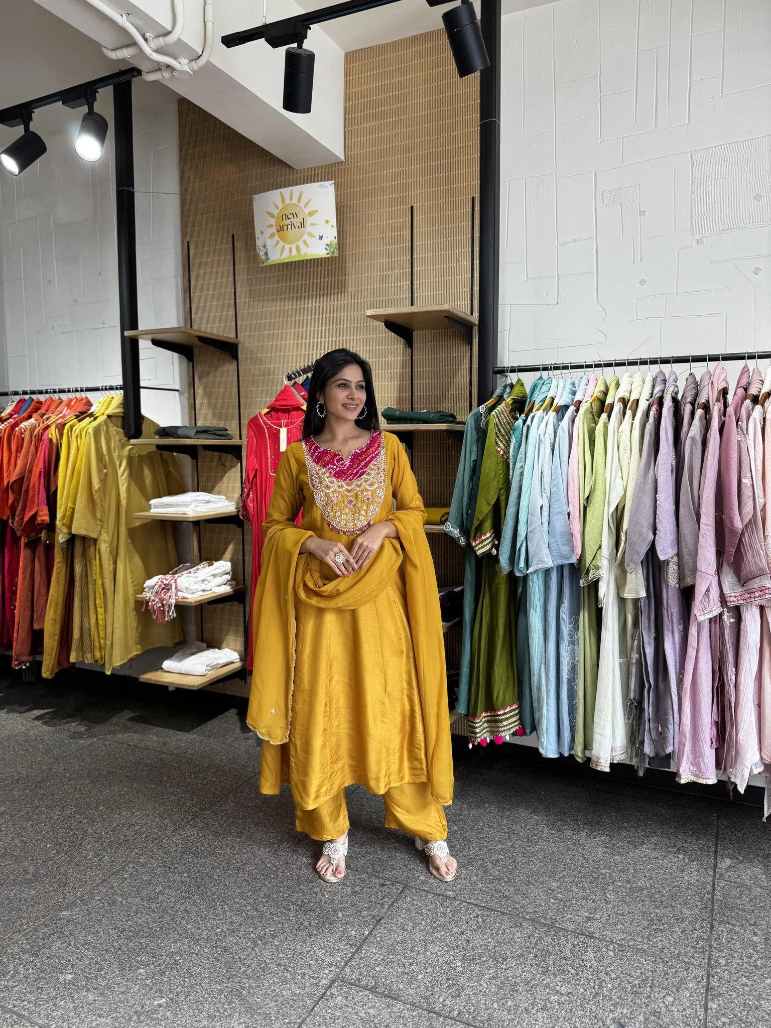 A woman modeling a mustard yellow traditional Indian outfit with a pink embroidered neckline and a matching dupatta, standing inside a clothing store with racks of colorful garments in the background.