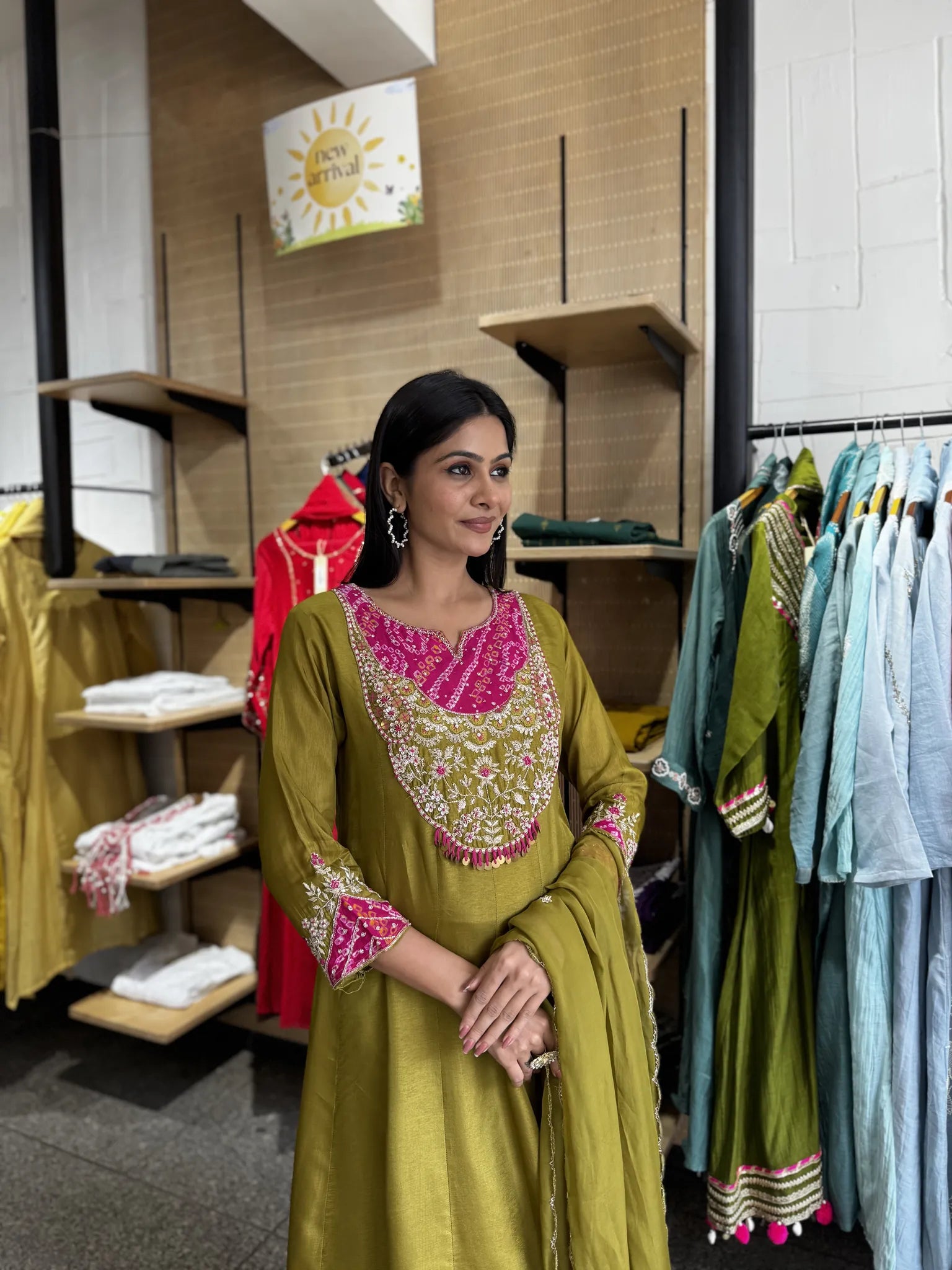 A woman modeling an olive green traditional Indian salwar kameez suit with vibrant pink and gold embroidery, standing inside a clothing store.