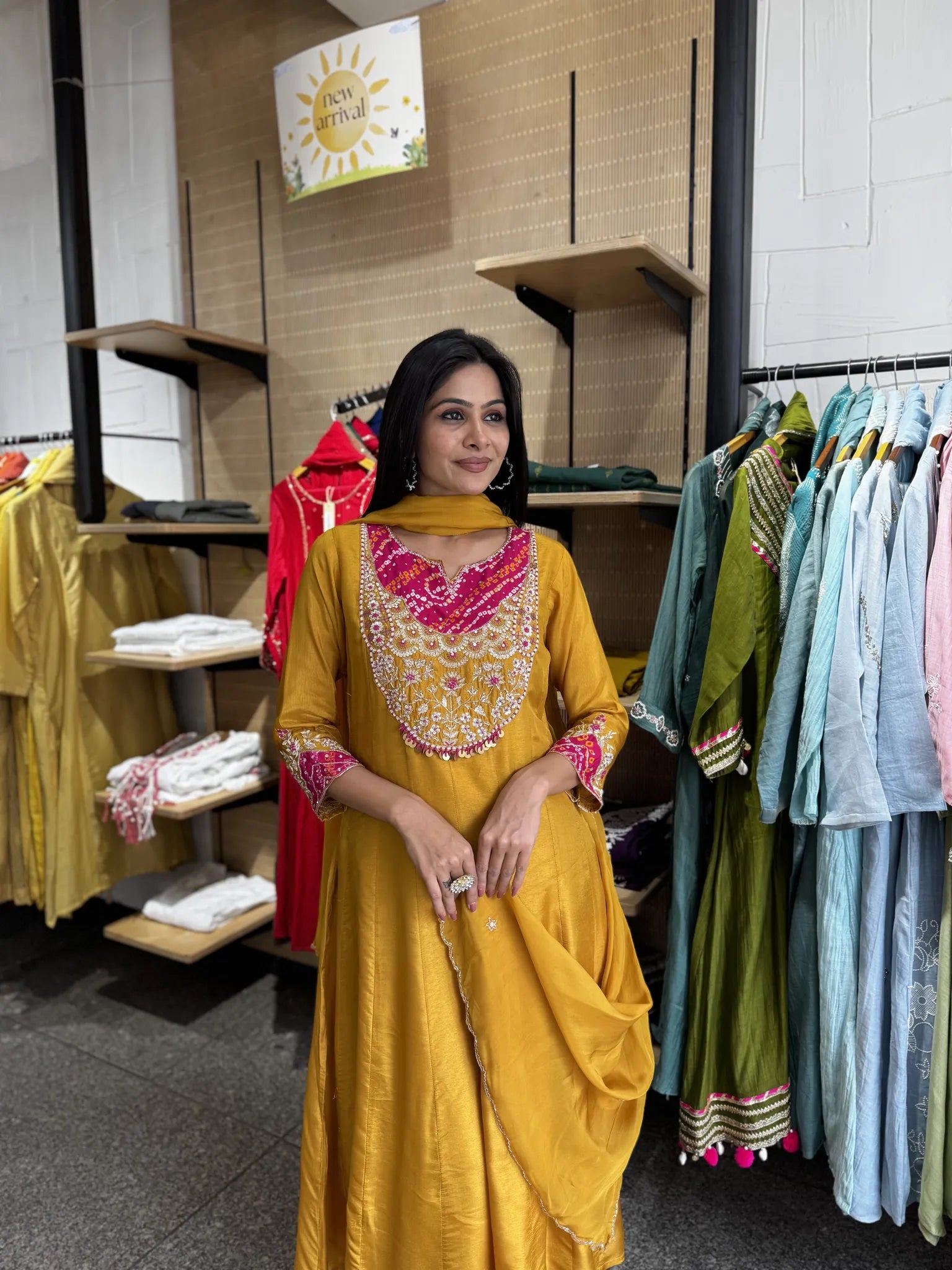 A woman wearing a mustard yellow kurta with pink and gold embroidery stands inside a clothing store, with racks of other garments visible in the background.