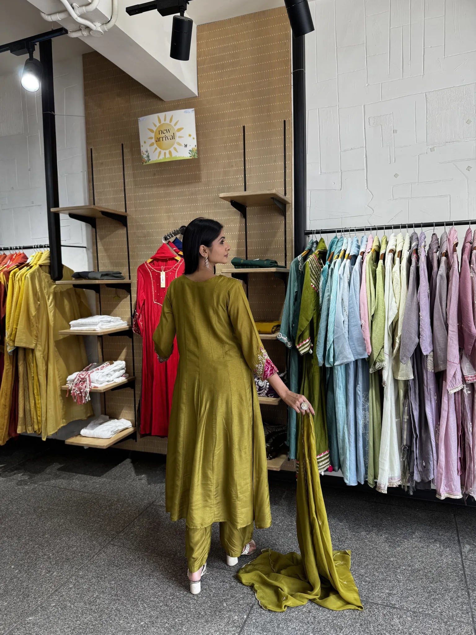 A woman stands in a clothing store wearing a mustard yellow kurta and matching pants, holding a matching dupatta. Clothing racks with various colors of garments are visible on both sides.