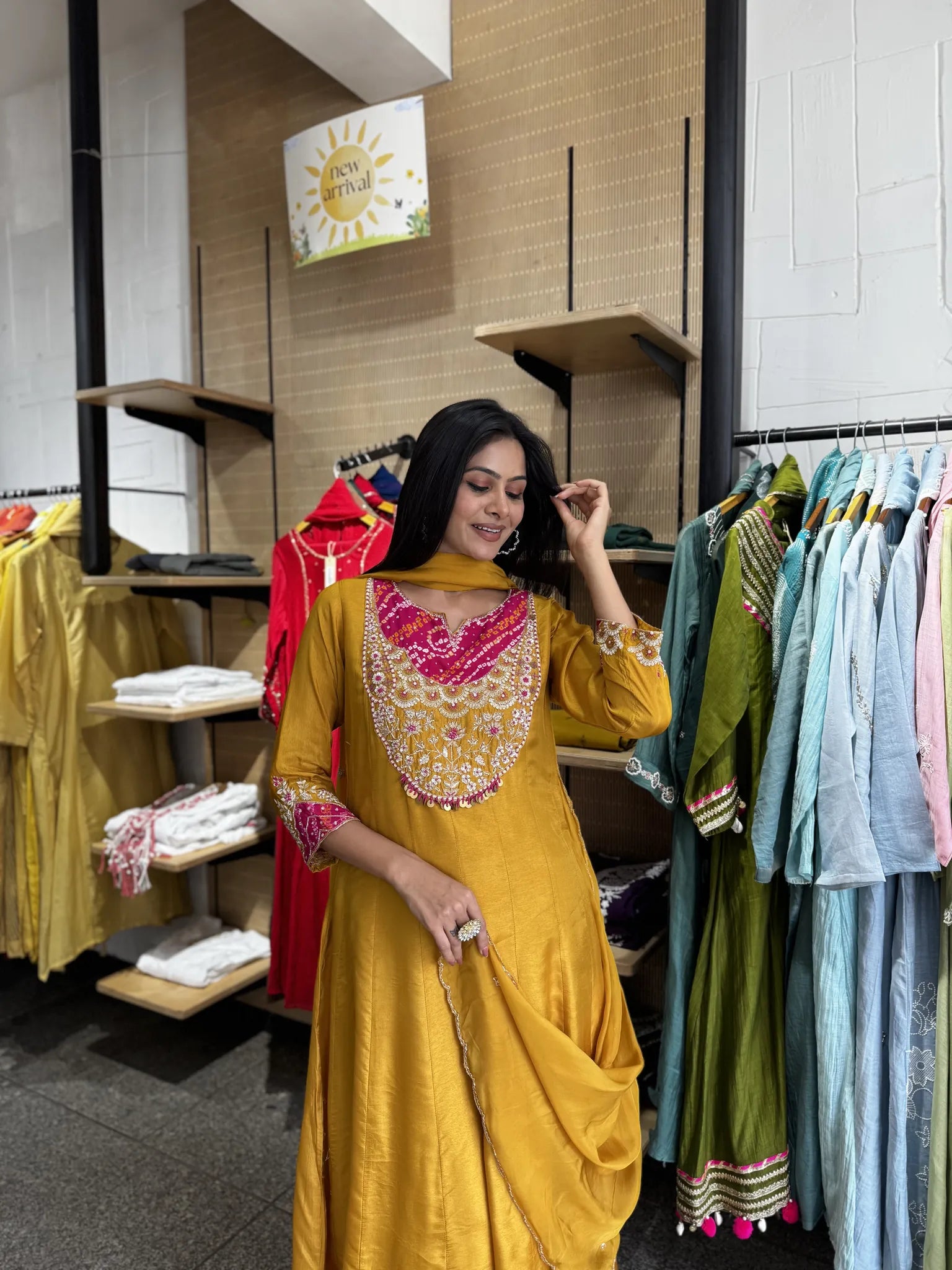 A woman modeling a mustard yellow traditional Indian salwar kameez set with a heavily embroidered pink and gold neckline, standing inside a clothing store.