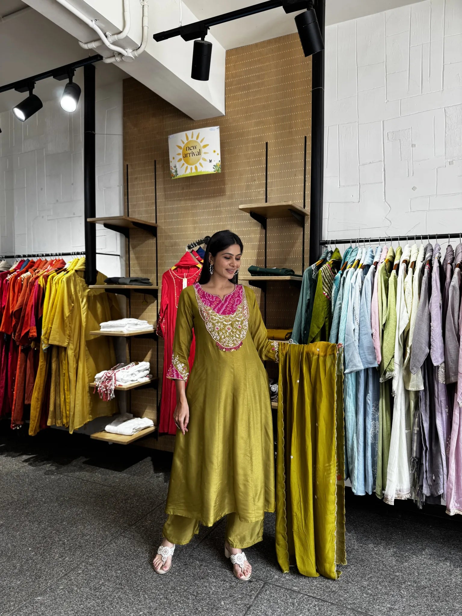 A woman modeling a mustard yellow traditional Indian outfit with a bright pink embroidered neckline, standing in a clothing store with racks of colorful garments in the background.