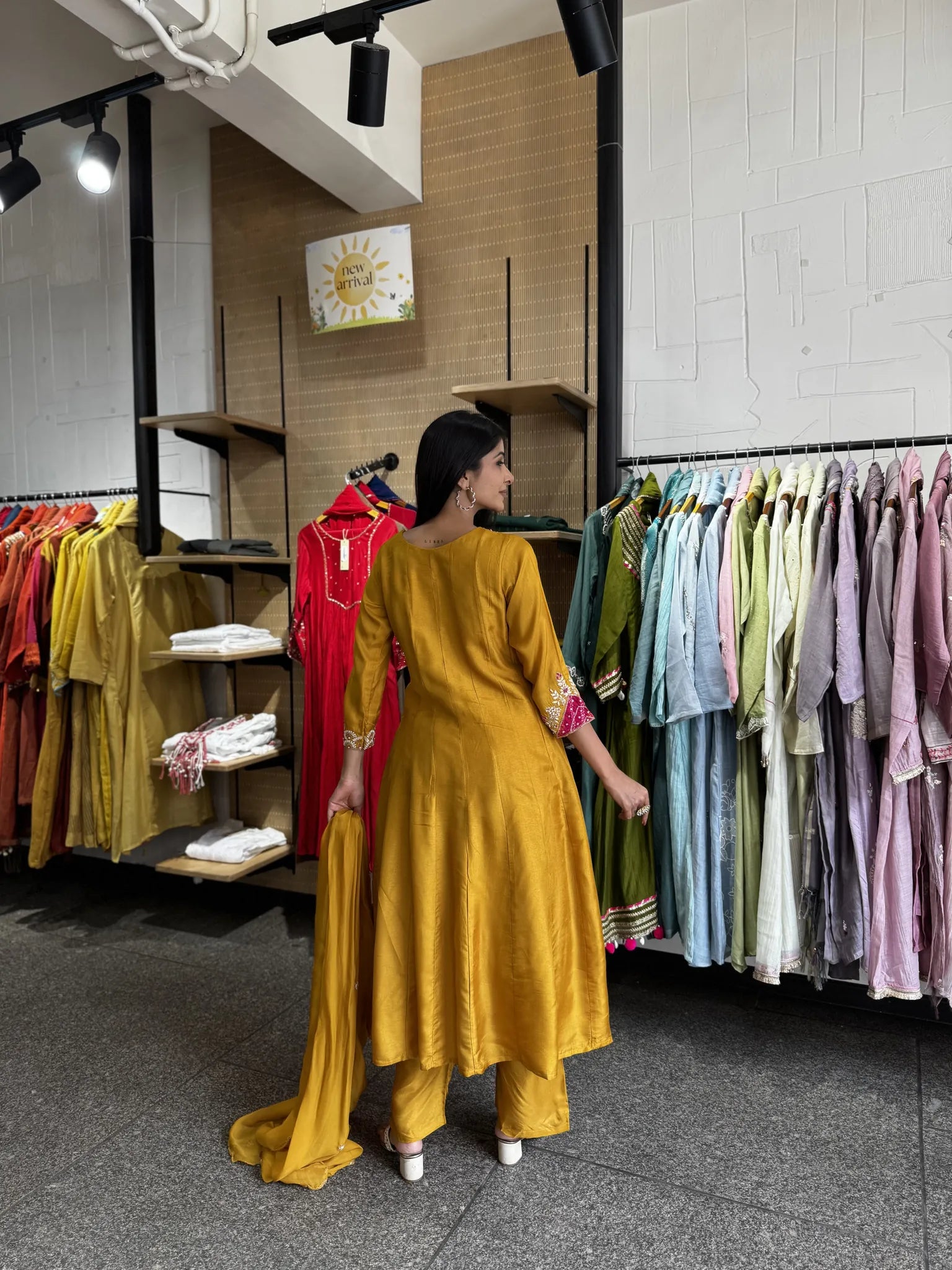 A woman wearing a mustard yellow traditional outfit stands in a clothing store, facing away from the camera. The store has racks of various colorful garments on both sides and a wood-paneled wall in the background.