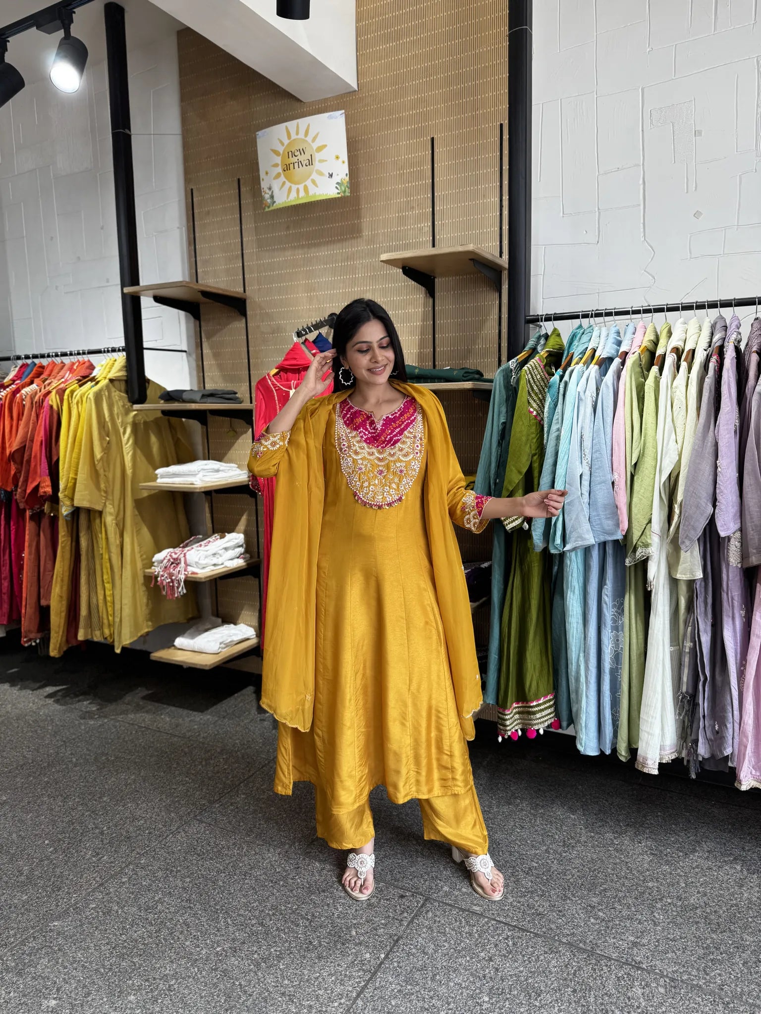 A woman modeling a bright yellow traditional Indian outfit with a matching draped outer layer and a heavily embroidered pink and gold neckline, standing inside a clothing store with racks of garments in the background.