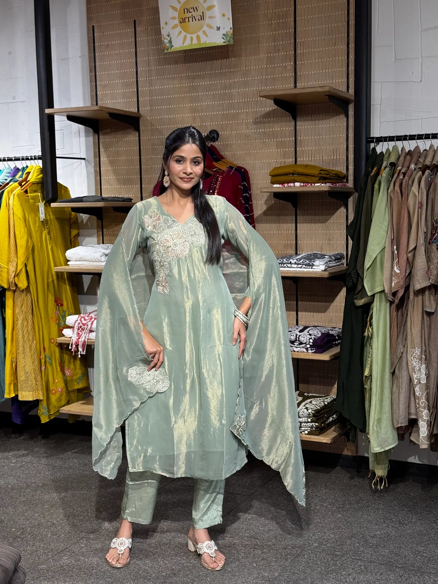 A woman modeling a mint green tissue A-line kurta pant set with a sheer matching dupatta, standing inside a clothing boutique with racks of garments behind her.