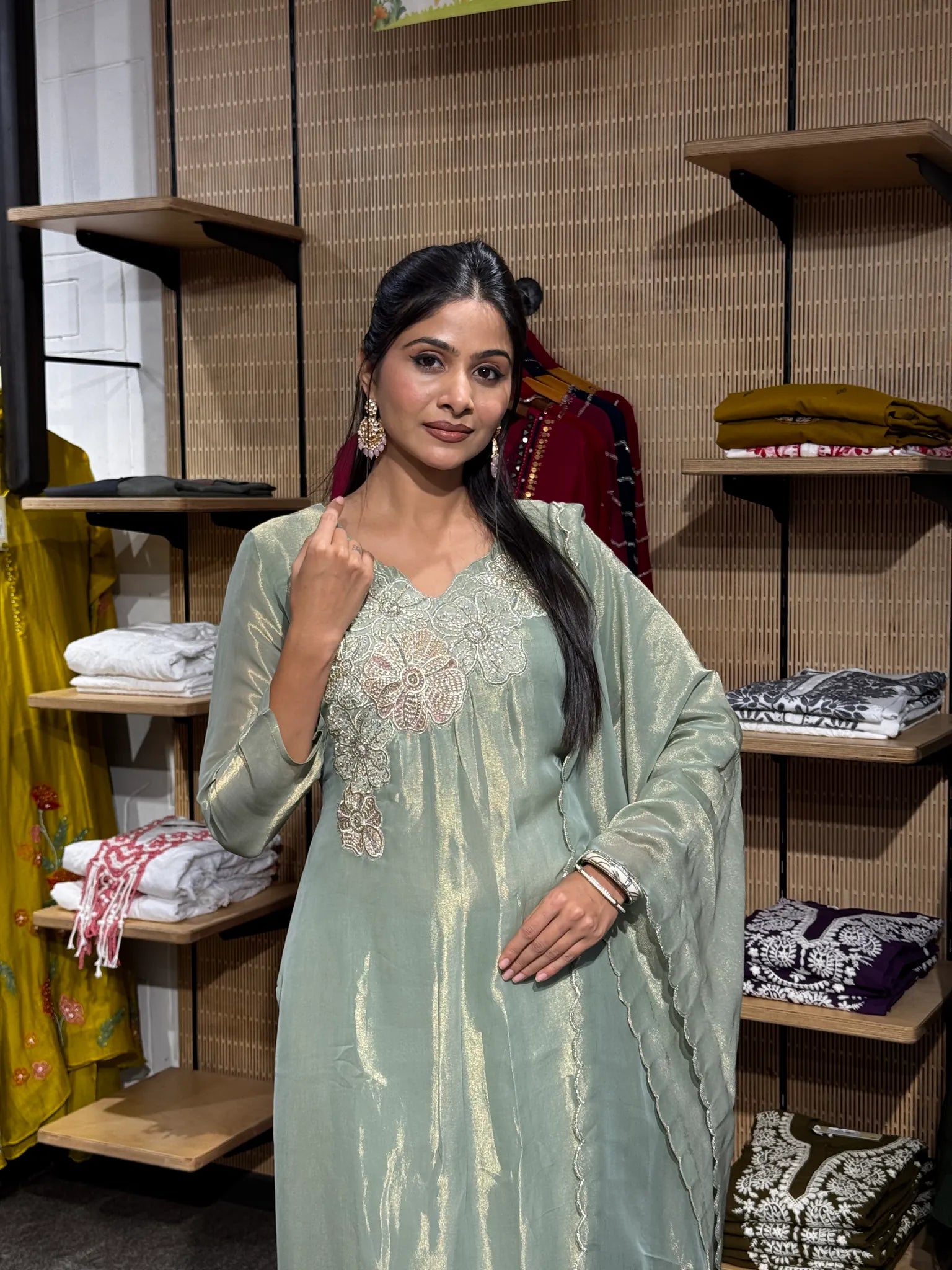 A woman wearing a mint green tissue A-line kurta with an embroidered neckline, holding the collar, inside a clothing store with folded fabrics on the shelves behind.