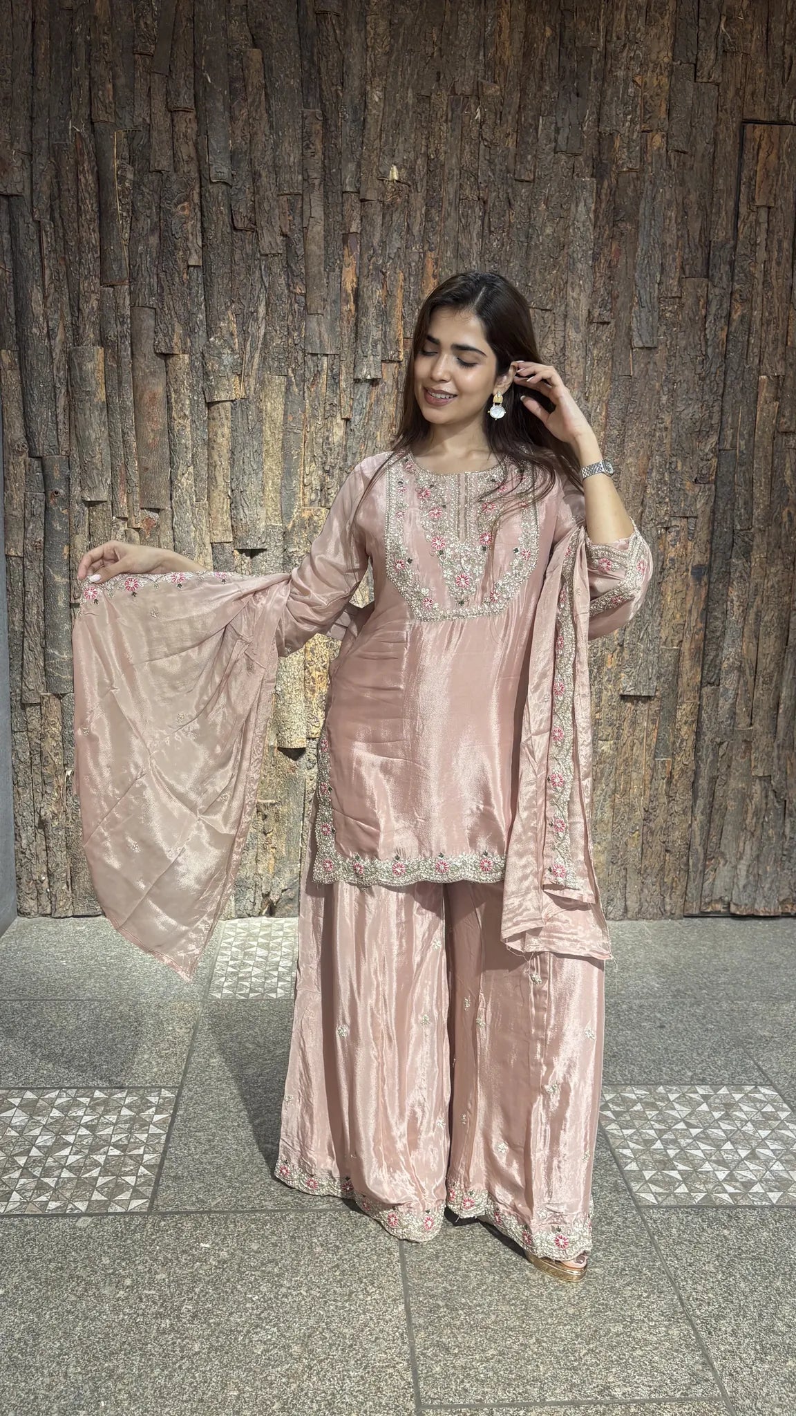 Onion pink chinnon short kurta with a wide plazo, the matching dupatta held open to one side against a timber wall.