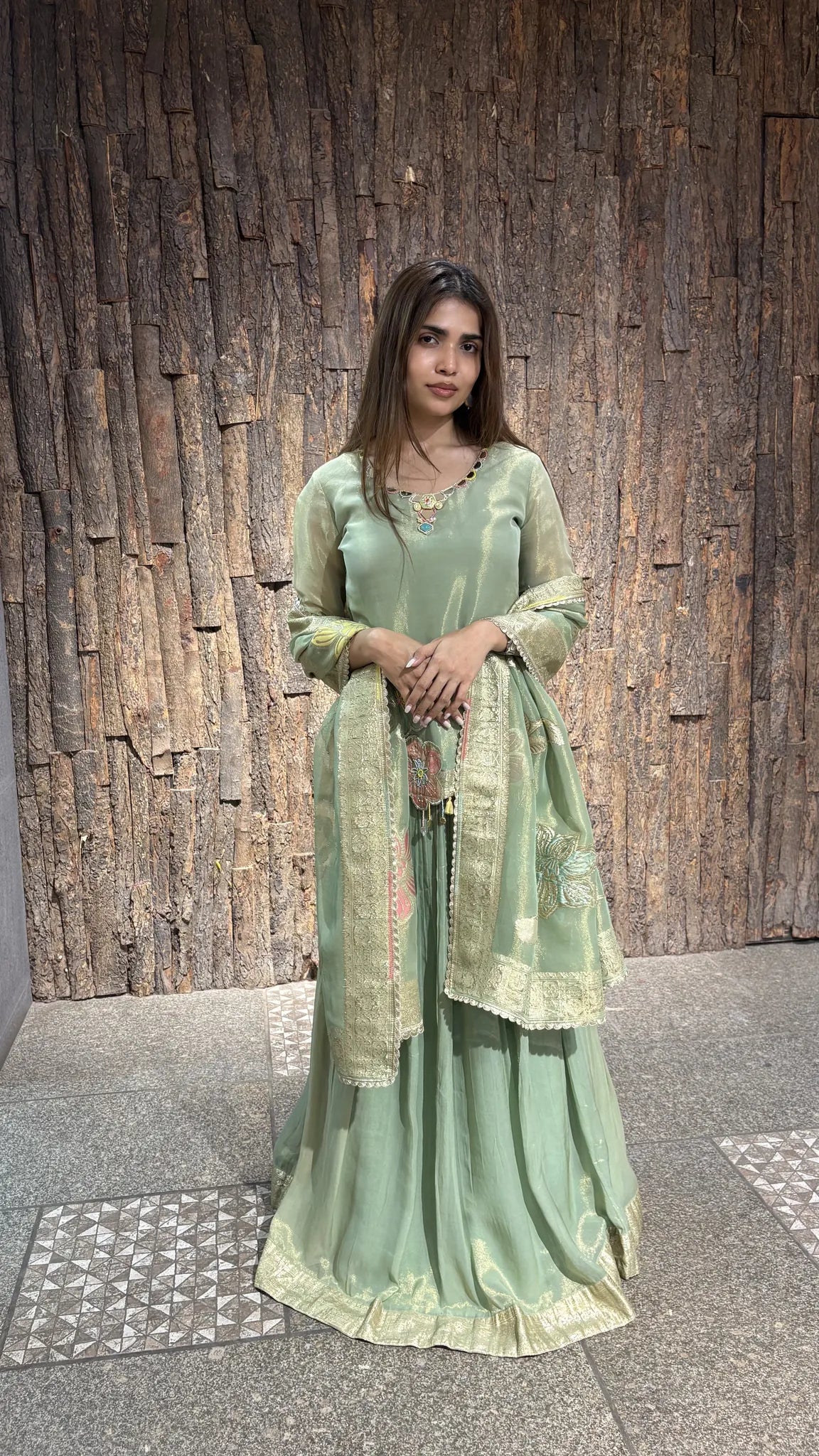 Pista green tissue kurta over a full gathered skirt, gold zari dupatta drawn over one shoulder, photographed against a textured timber wall.