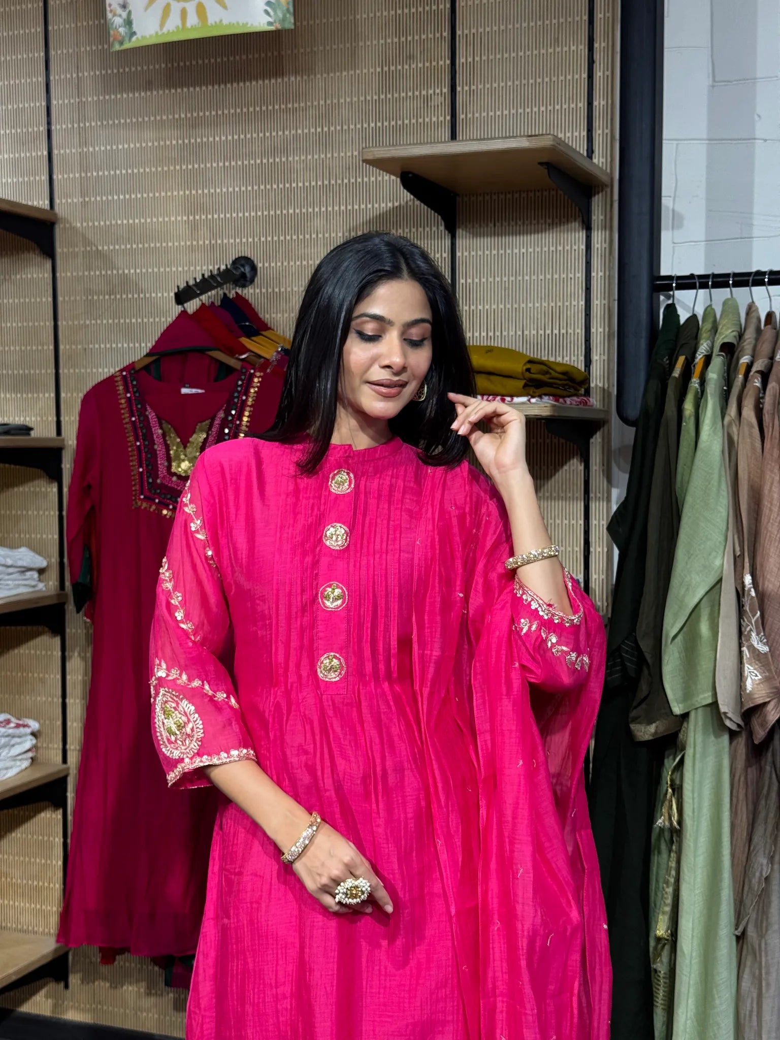 A woman wearing a rani pink mul chanderi A-line kurta with a stand collar and gold button placket, inside a clothing store.