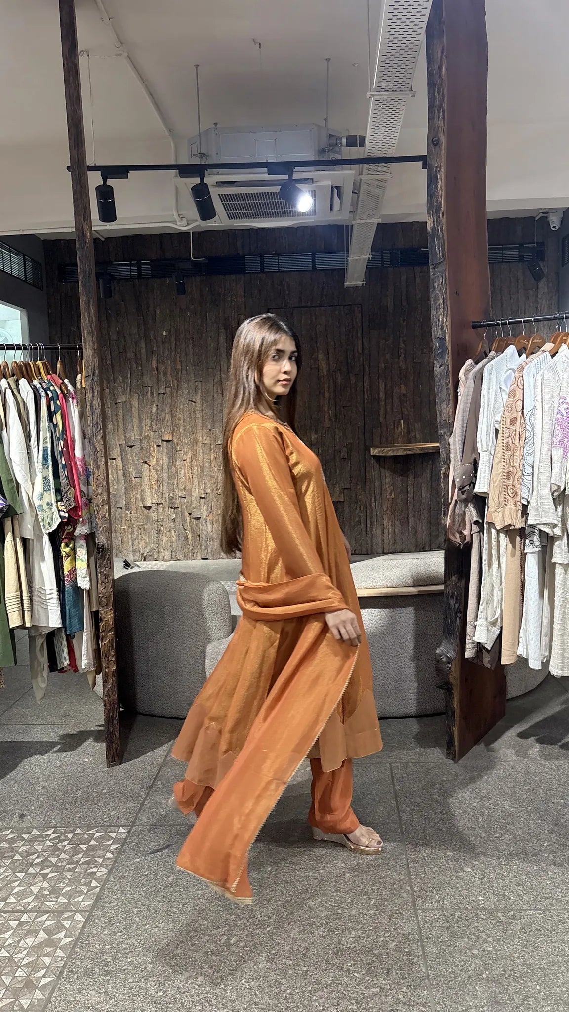 Rust orange tissue A-line kurta set seen in profile mid-walk, the long matching dupatta sweeping behind the pant.