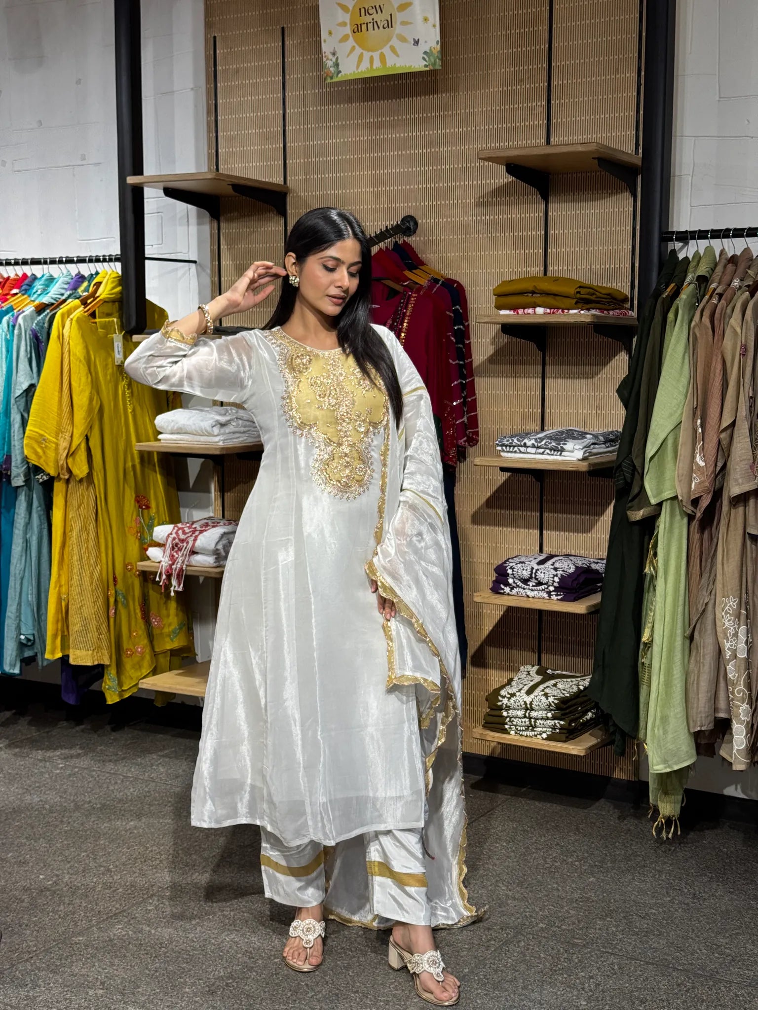 A woman modeling a white tissue round neck A-line kurta pant set with a sheer dupatta, standing inside a clothing boutique.