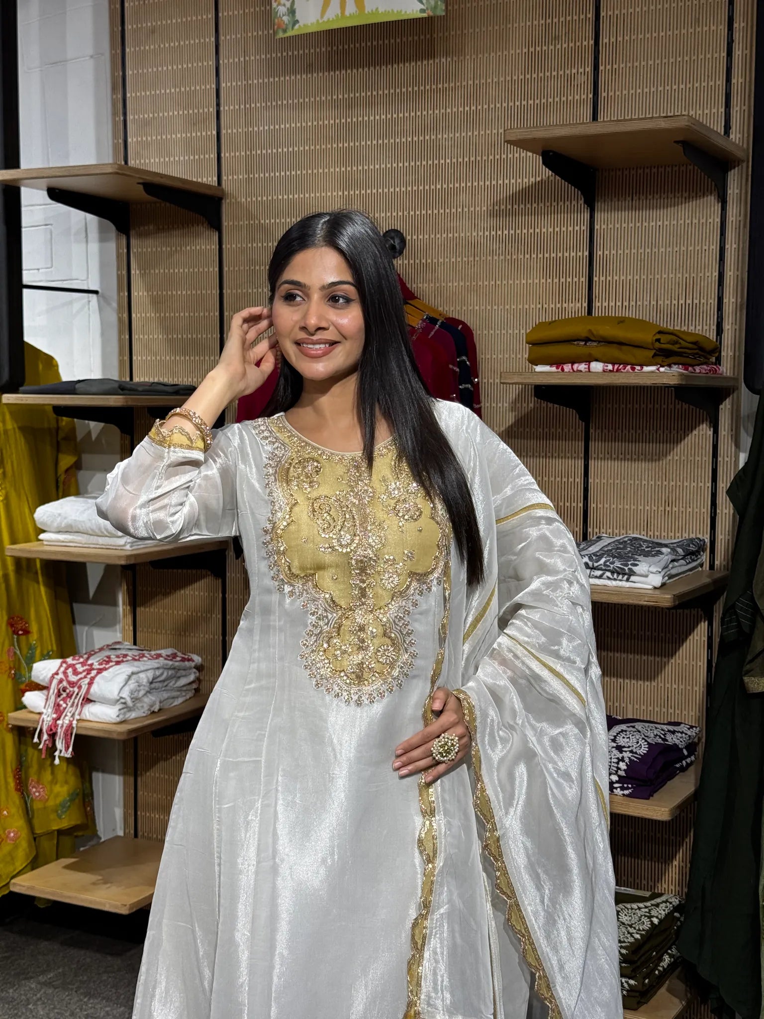 A woman in a white tissue kurta with a gold zari and sequin embroidered yoke, one hand raised, inside a clothing store.